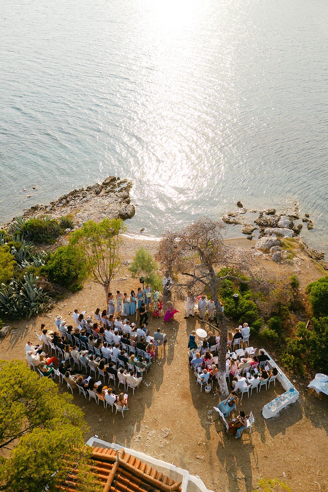 Aerial view of an outdoor wedding ceremony on a coastal cliff overlooking the ocean with guests seated in white chairs at Poros