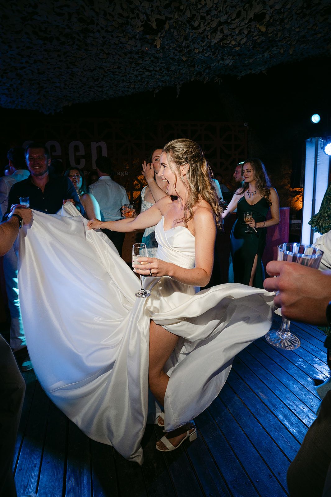 A person in a white wedding dress dances energetically on a dark dance floor while holding a wine glass at Poros