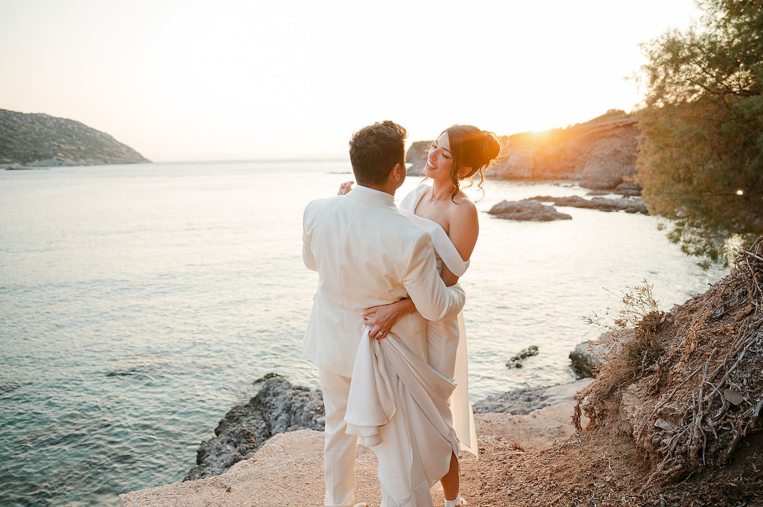Couple embraces on rocky coastal cliff during golden sunset over calm ocean waters at Ktima 48 Athens