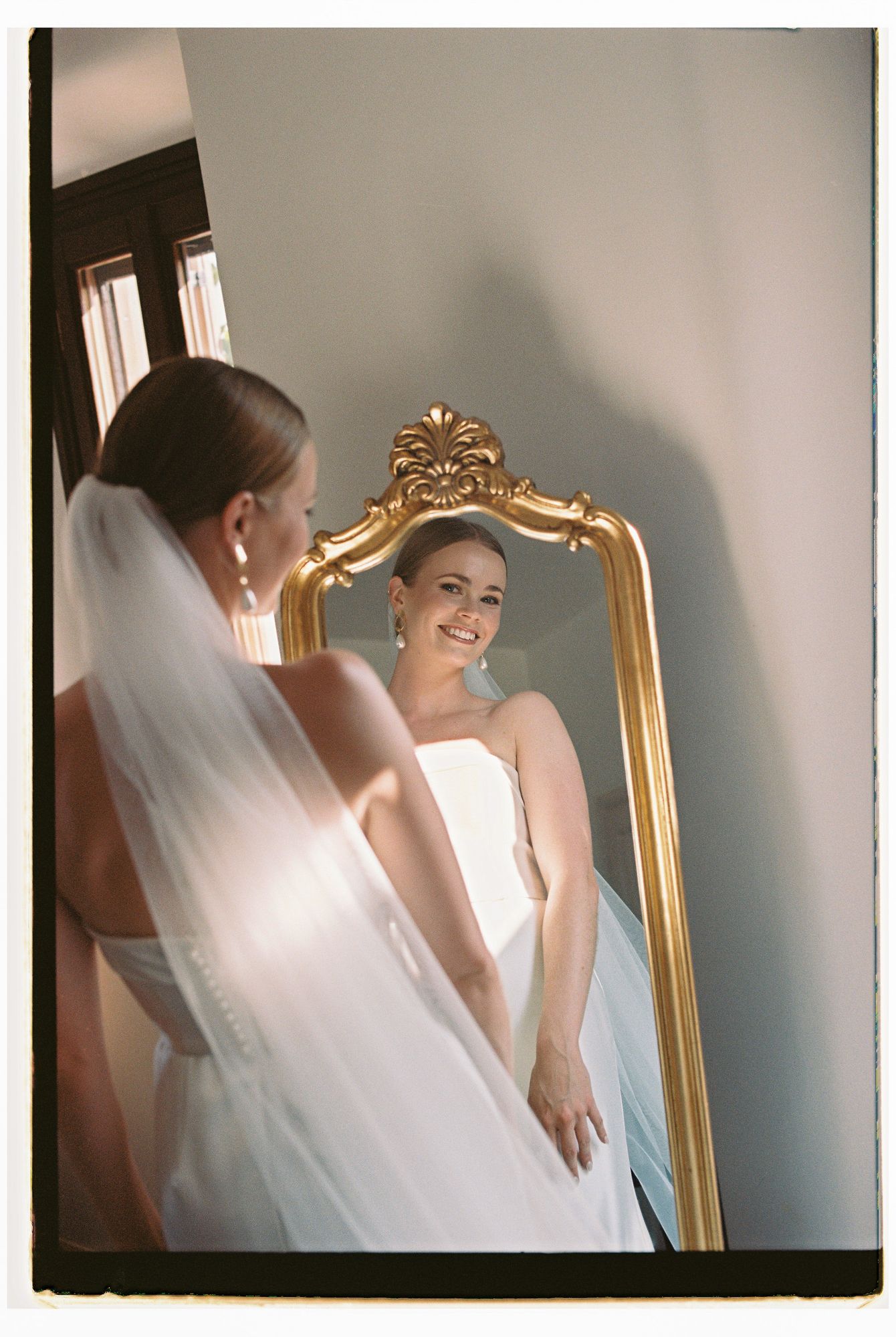 A bride in a white wedding dress admires her reflection in an ornate gold-framed mirror.