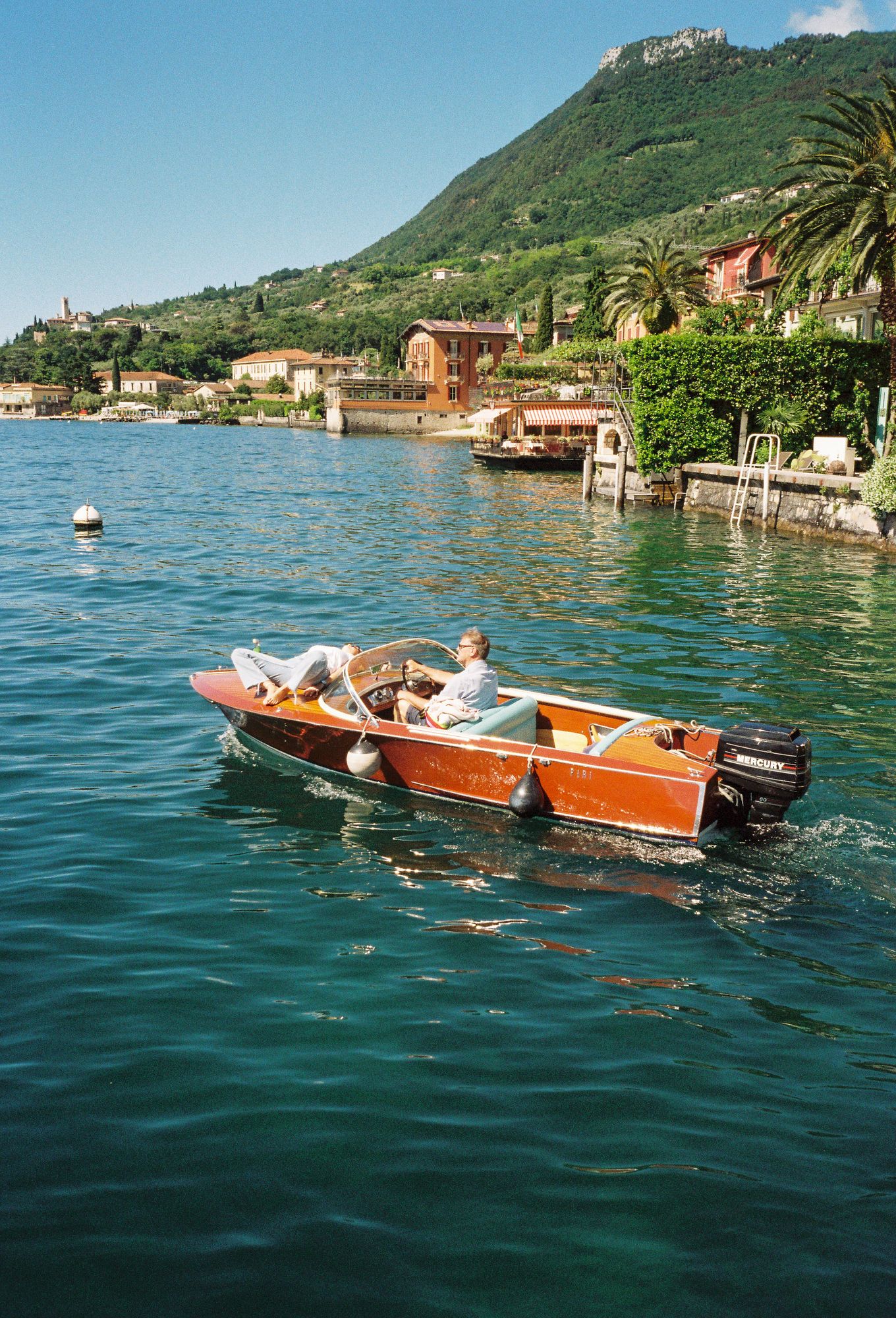 Wooden vintage boat cruising on blue Mediterranean waters near mountainous coastline with colorful buildings and villas at lake Garda