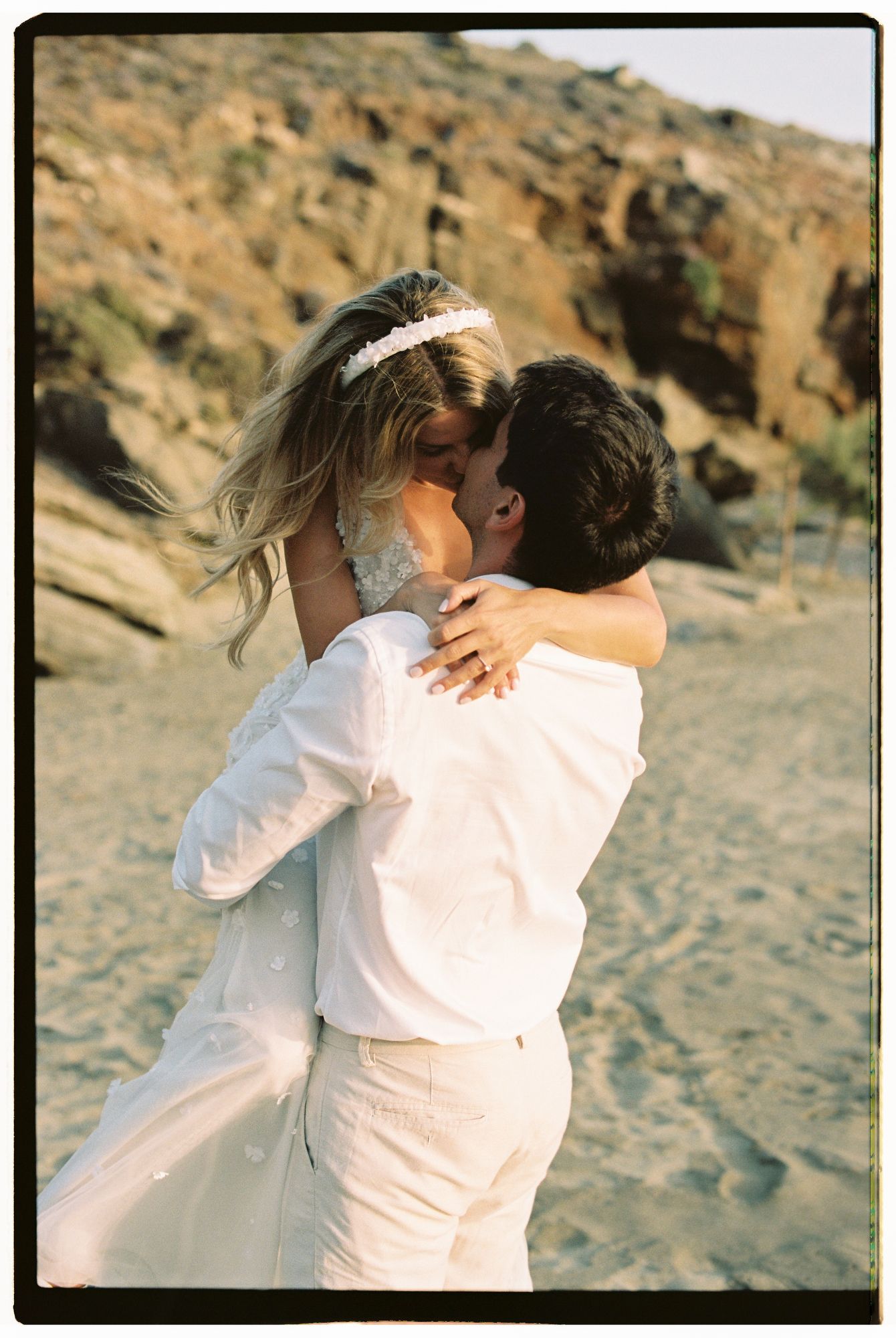 A couple in white outfits embraces tenderly on a sandy beach with rocky cliffs in the background.