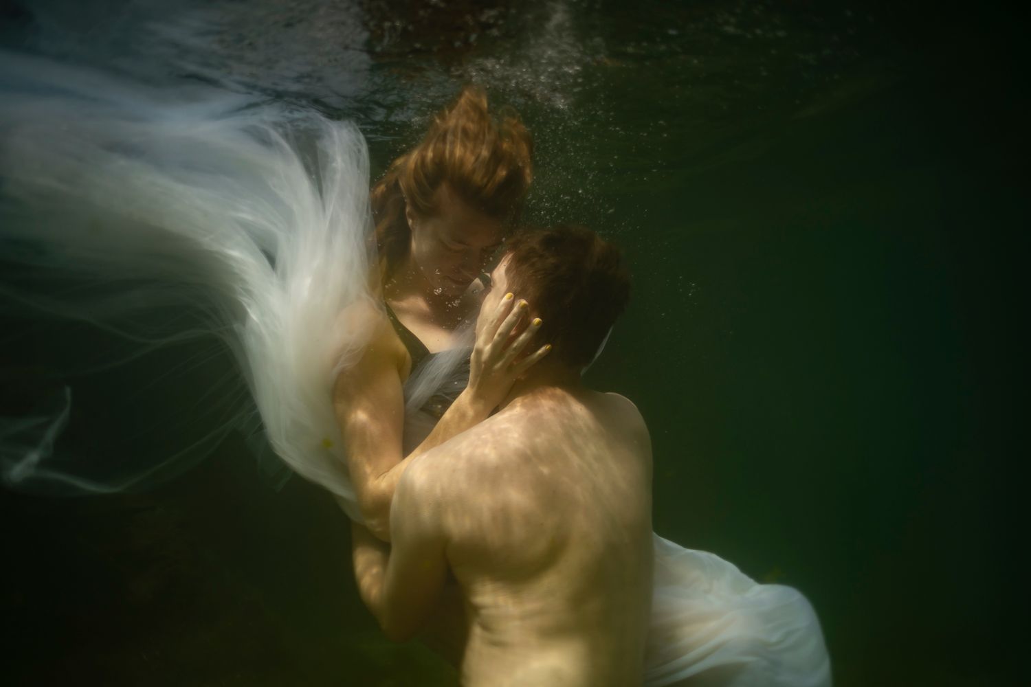 Couple sharing a romantic underwater kiss, bride in flowing white dress, ethereal green water surroundings.