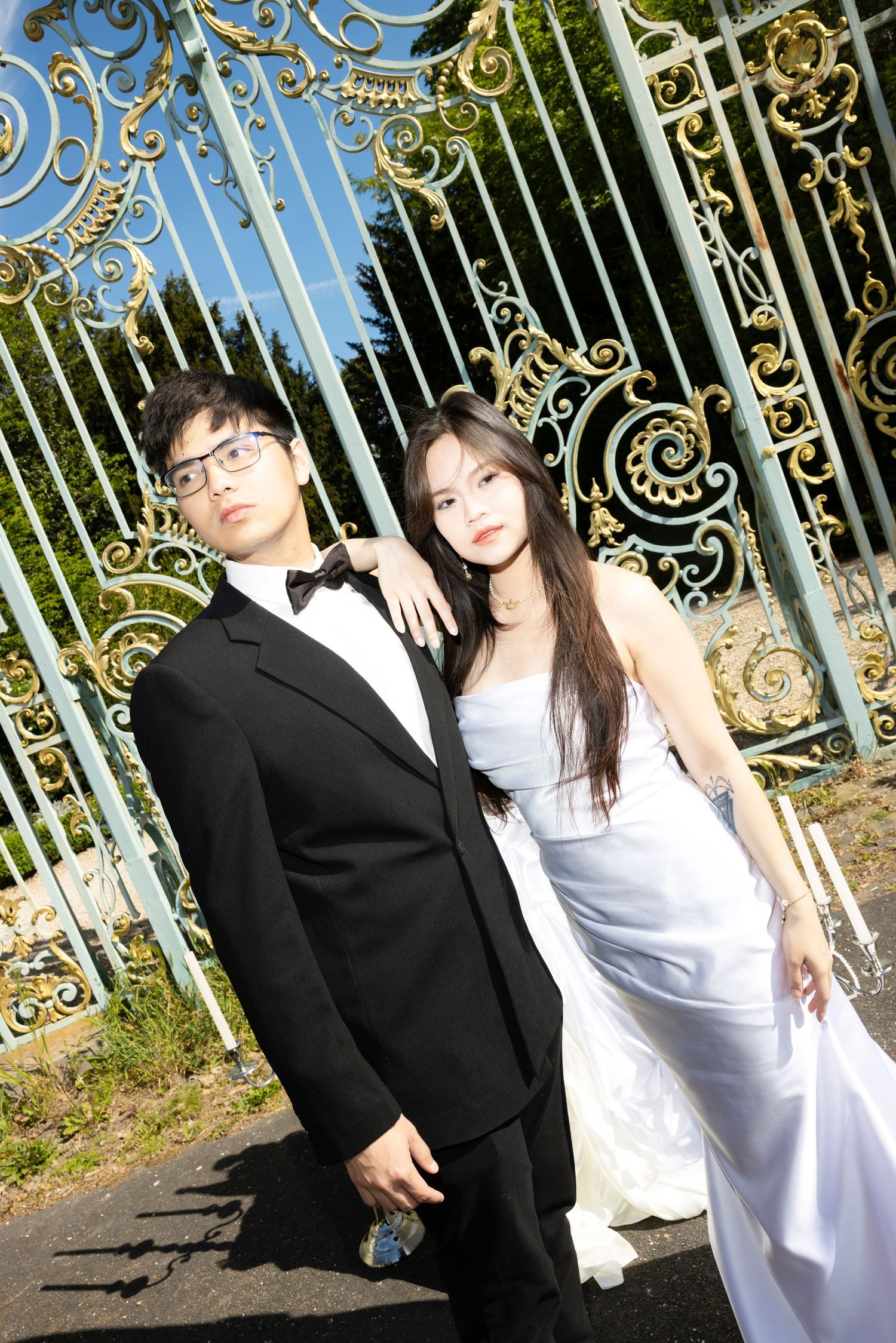 Asian couple in wedding attire posing before ornate gold and teal gates, groom in tuxedo, bride in white gown.