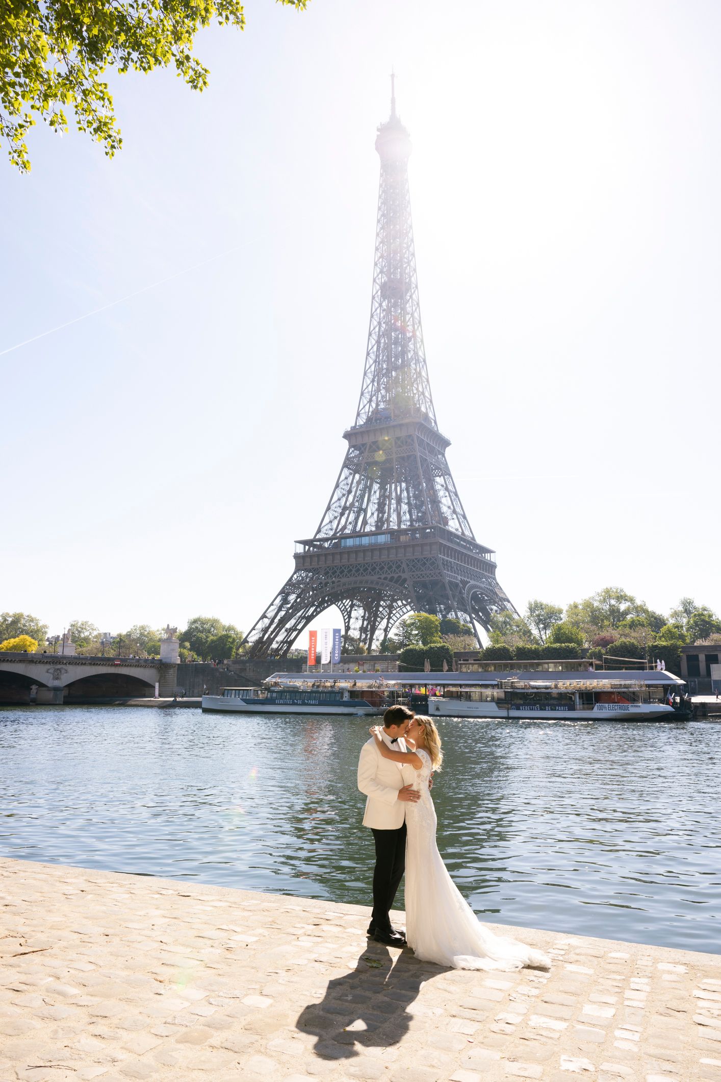 Bride and groom embrace in front of the Eiffel Tower beside the Seine River on a sunny day in Paris.