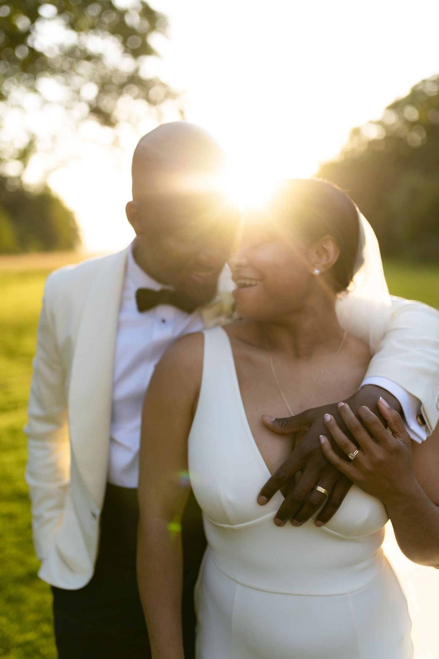 A joyful Black couple in wedding attire embraces during a golden sunset on a lush green lawn.