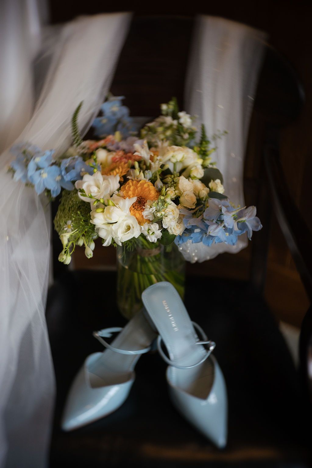 Blue bridal heels beside a pastel wedding bouquet with hydrangeas and roses in a glass vase.