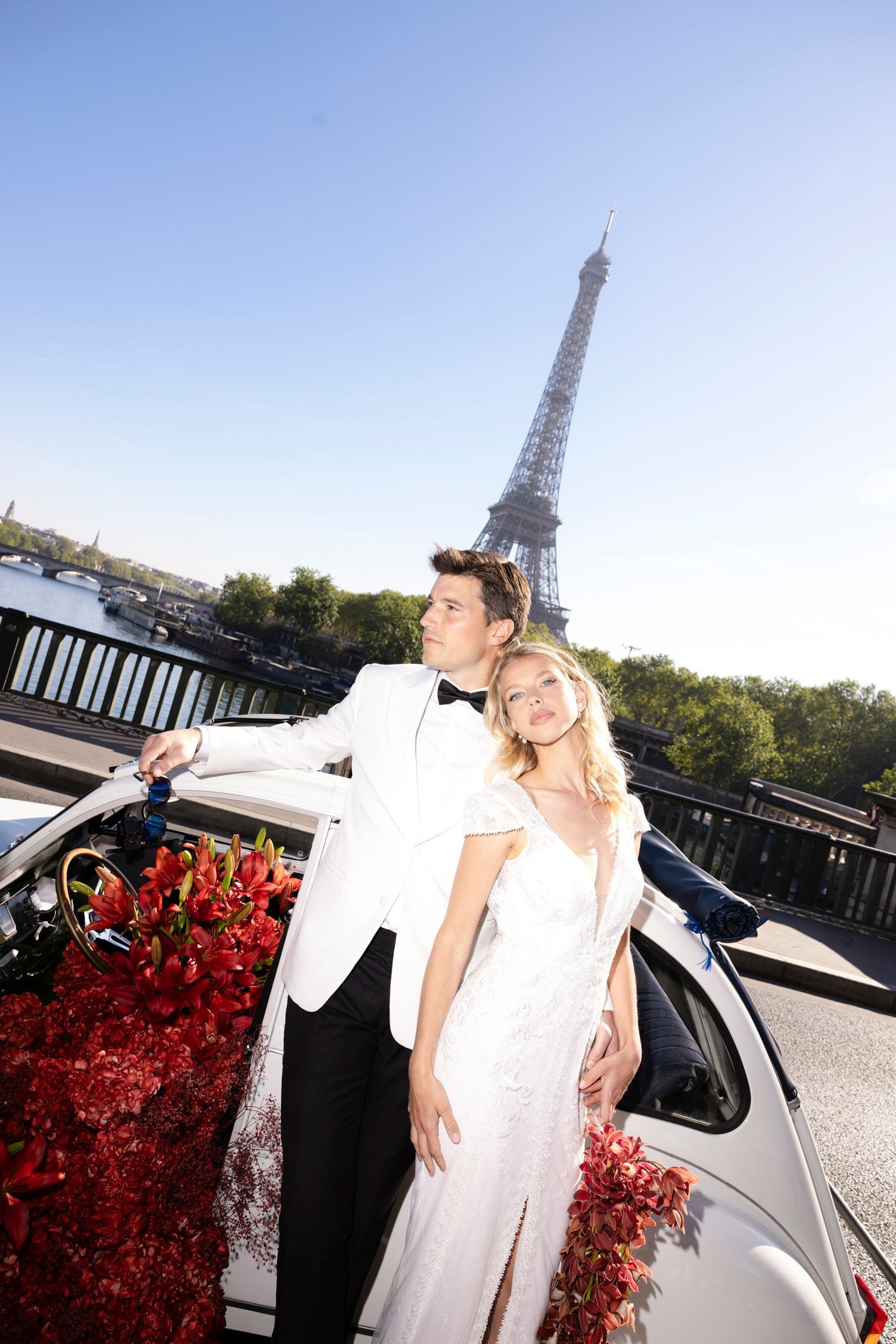 Newlywed couple posing with a flower-adorned car in front of the Eiffel Tower in Paris on a sunny day.