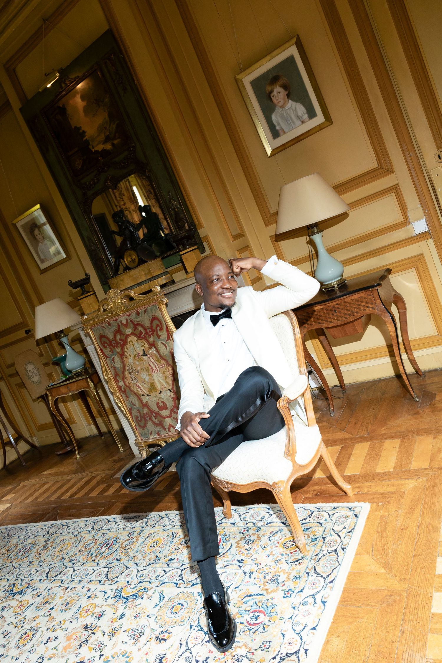 Elegant groom in white tuxedo jacket sitting in ornate chair in luxurious gold-paneled room with classic artwork.