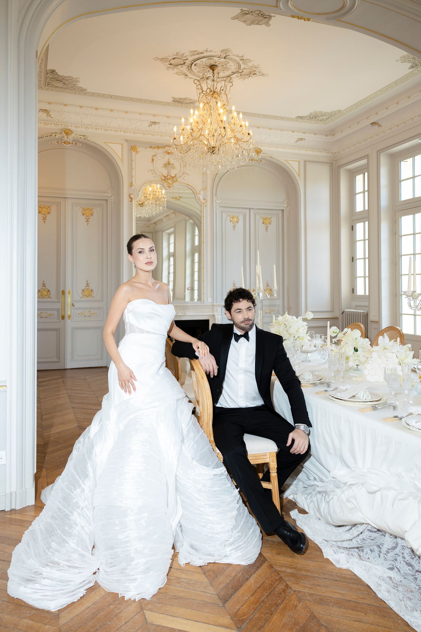 Elegant bride in white strapless gown and groom in black tuxedo posing in luxurious Parisian ballroom with chandelier.