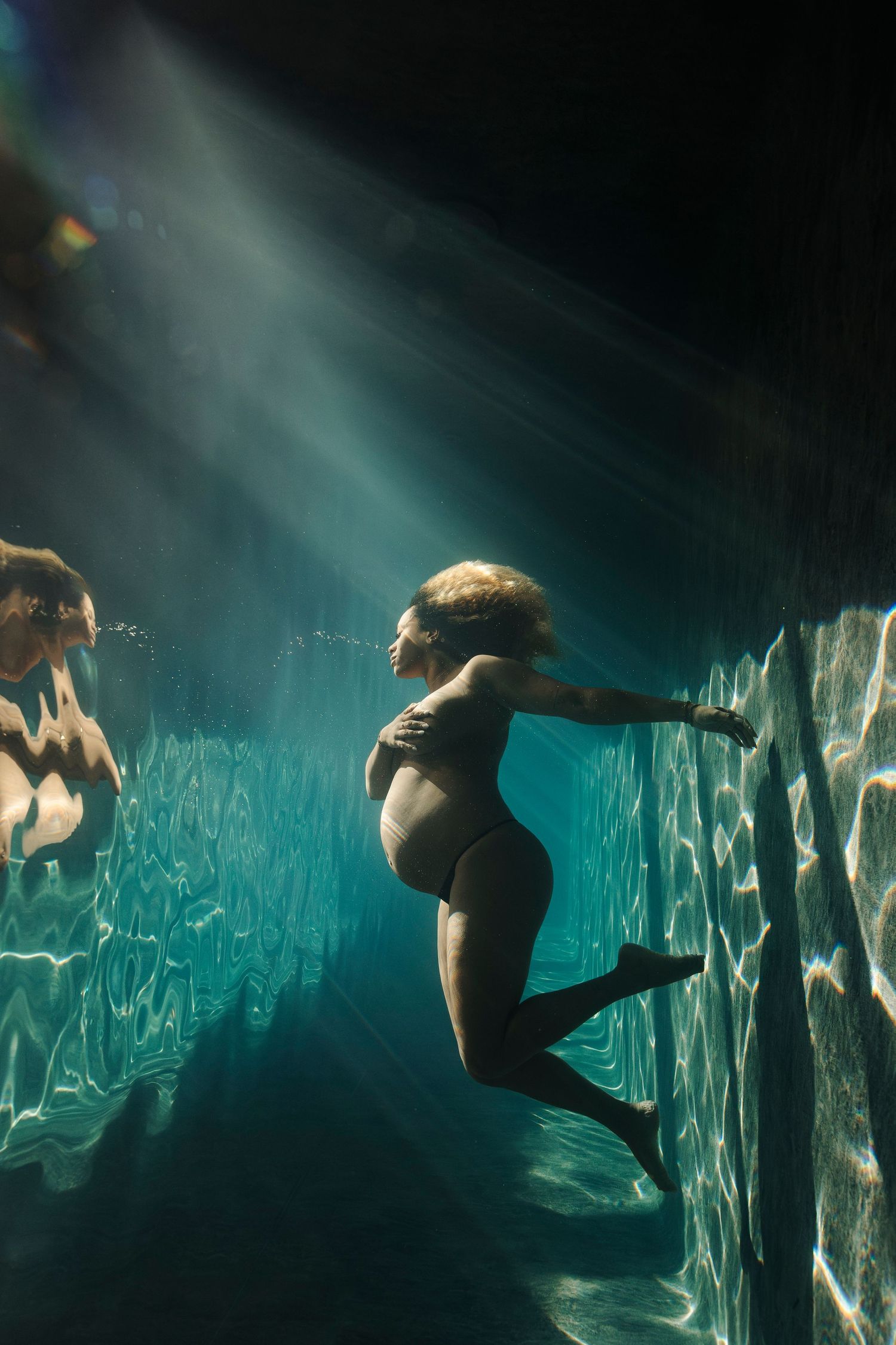 Pregnant woman swimming underwater in a pool with dramatic light rays filtering through the water surface.