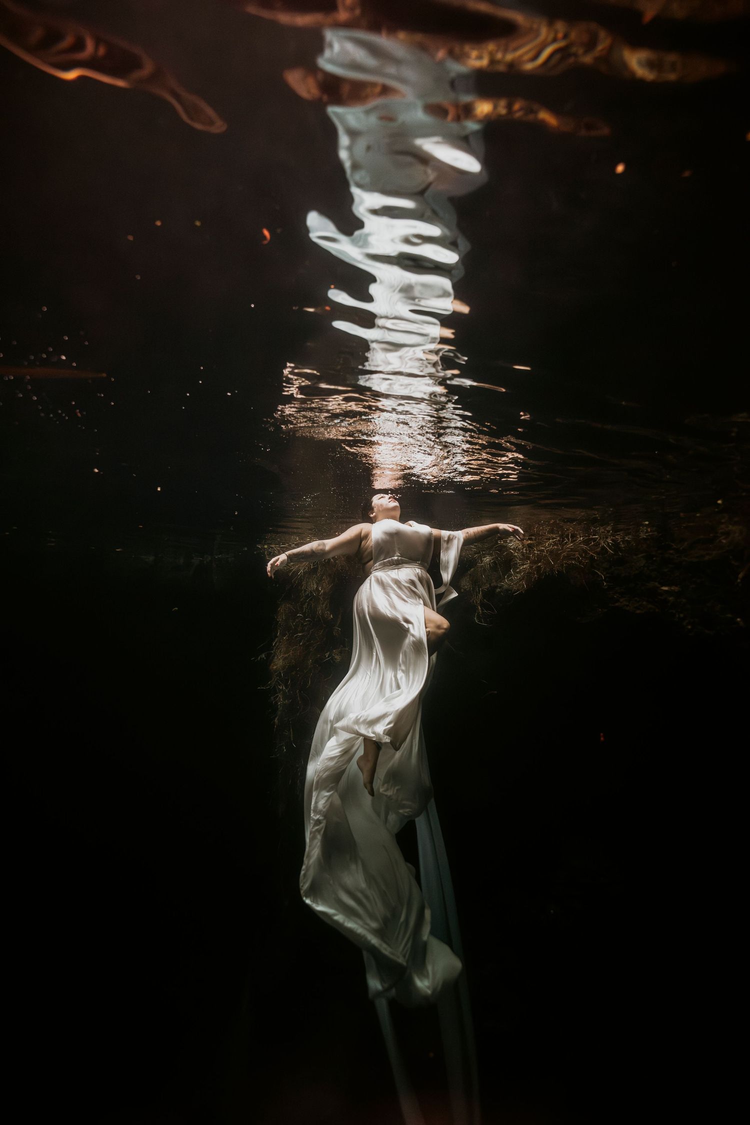 Ethereal underwater photo of a woman in a white flowing dress, arms outstretched, with light rippling on the surface above.