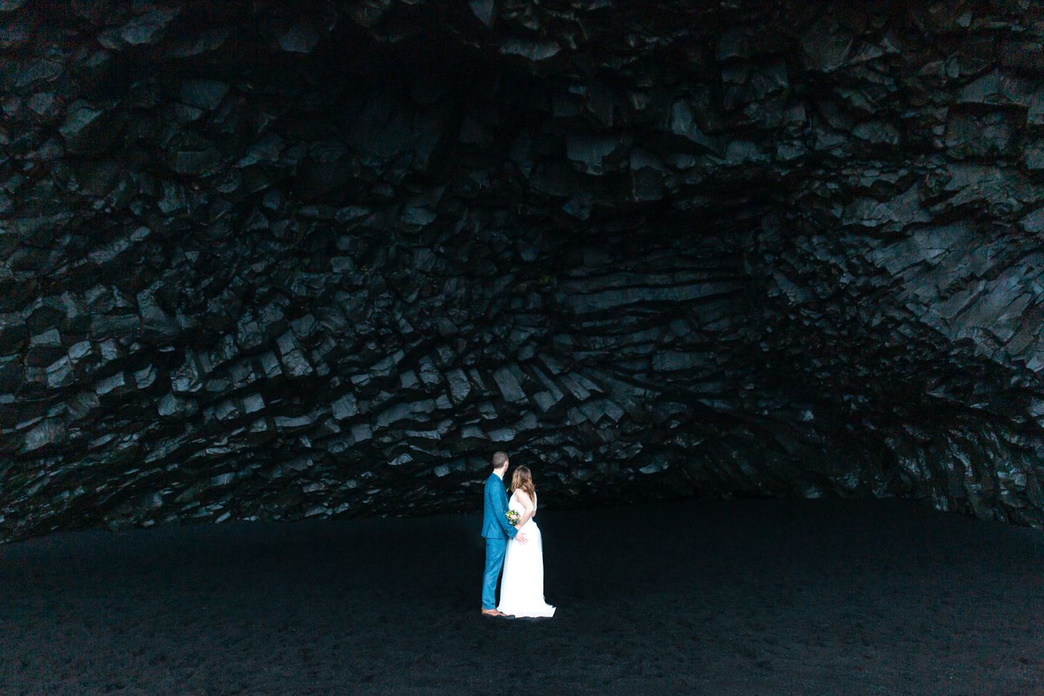 Bride and groom embracing on black sand beach beneath dramatic dark volcanic cave rock formation in Iceland.