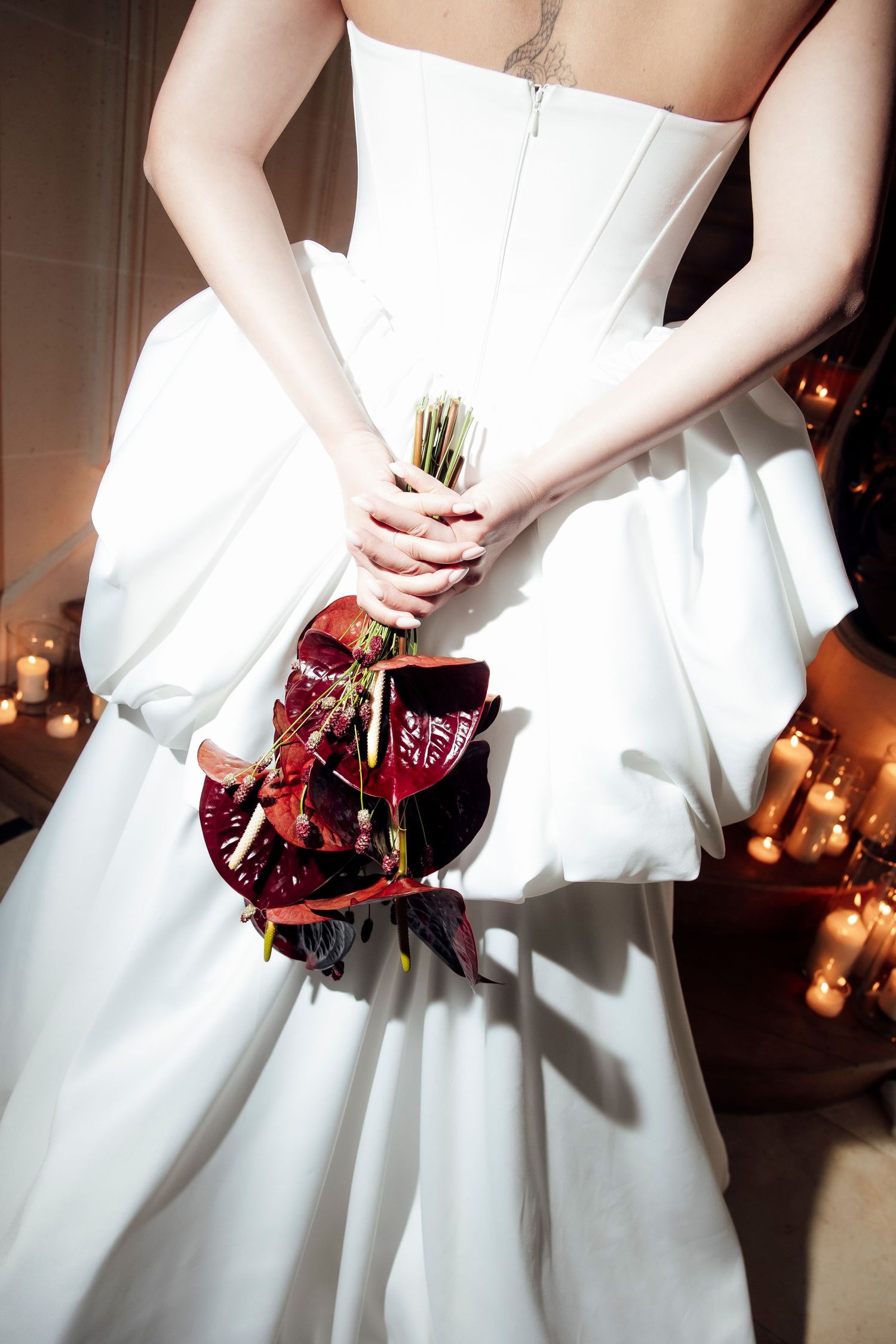 Bride in white strapless gown holding deep red floral bouquet behind her back, surrounded by candlelight.