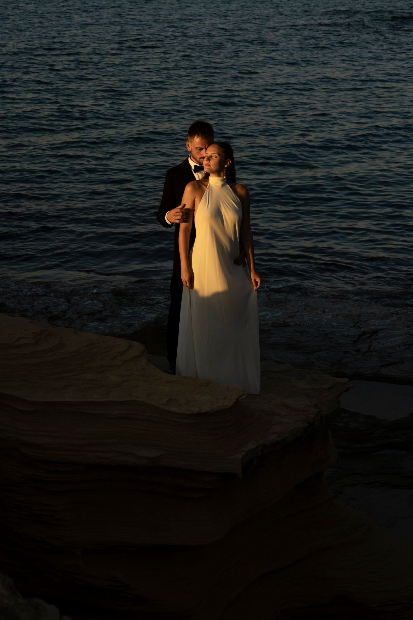 Couple in wedding attire embracing on rocky shore at sunset, bride in white gown, groom in dark suit, ocean backdrop.