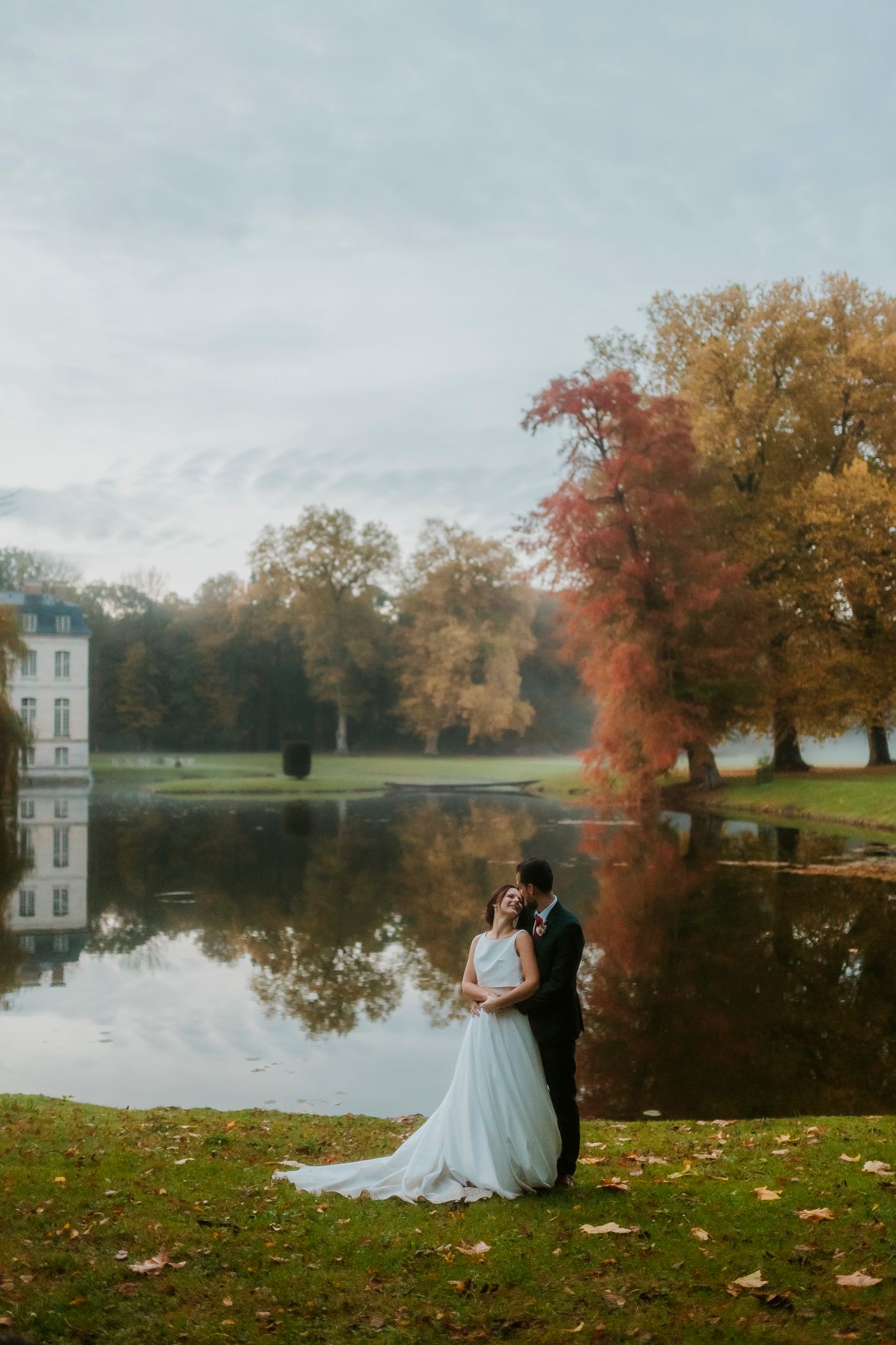 Bride and groom embrace by a reflective lake surrounded by autumn foliage near a French chateau. wedding photographer