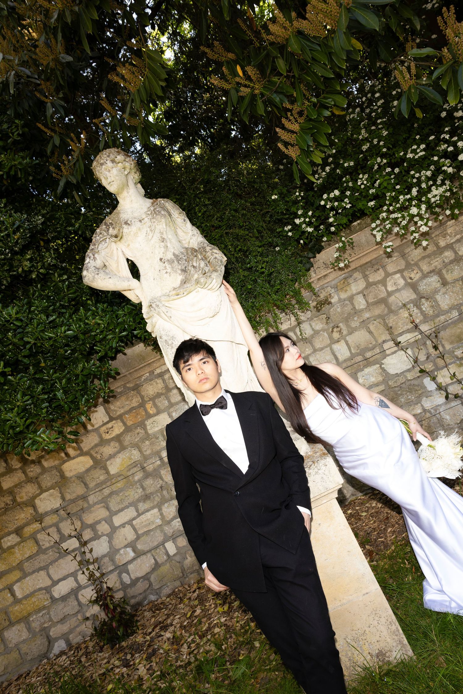 Asian bride and groom posing by a stone statue in a garden, wearing wedding attire with a brick wall backdrop.