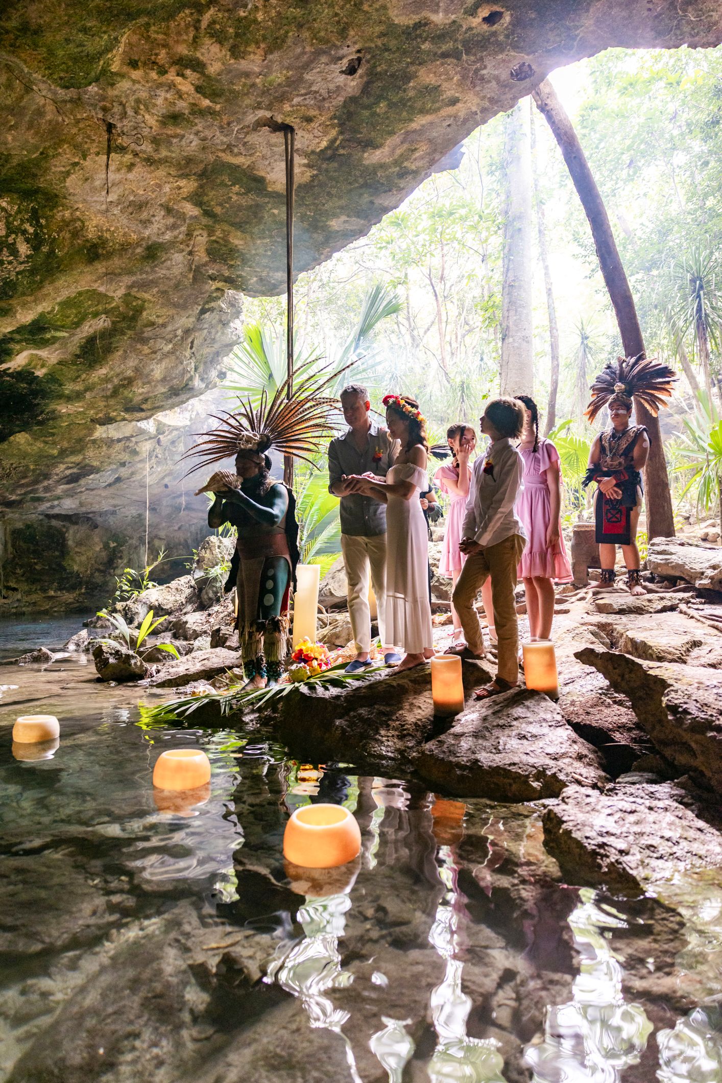 Mayan ceremony in a cenote cave with officiants in feathered headdresses, couple, and guests standing on rocks by water.