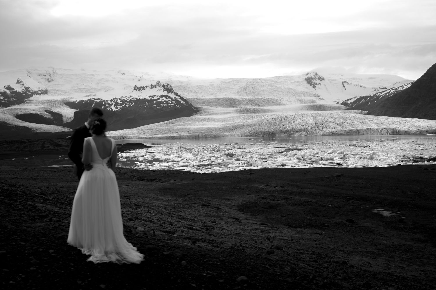 Black and white photo of a couple in wedding attire embracing before a vast glacier landscape in Iceland.