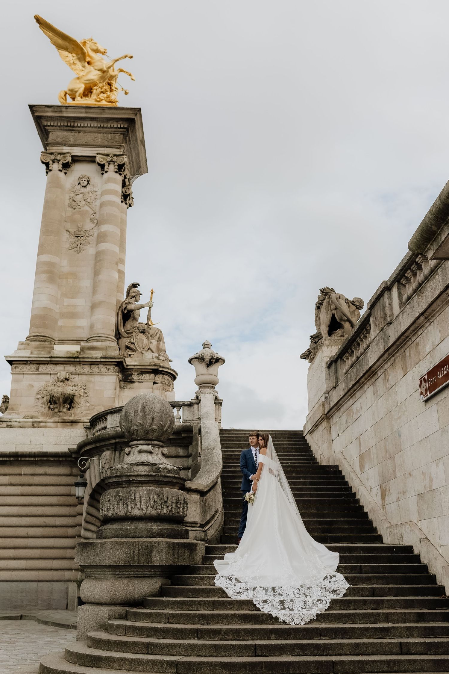 Bride and groom pose on ornate stone steps near a golden eagle statue at Pont Alexandre III, Paris.