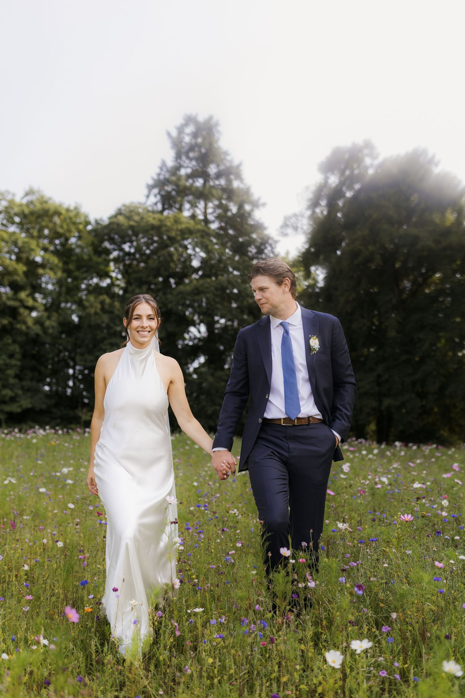 Bride and groom walk hand in hand through a wildflower meadow, she in white halter gown, he in navy suit.