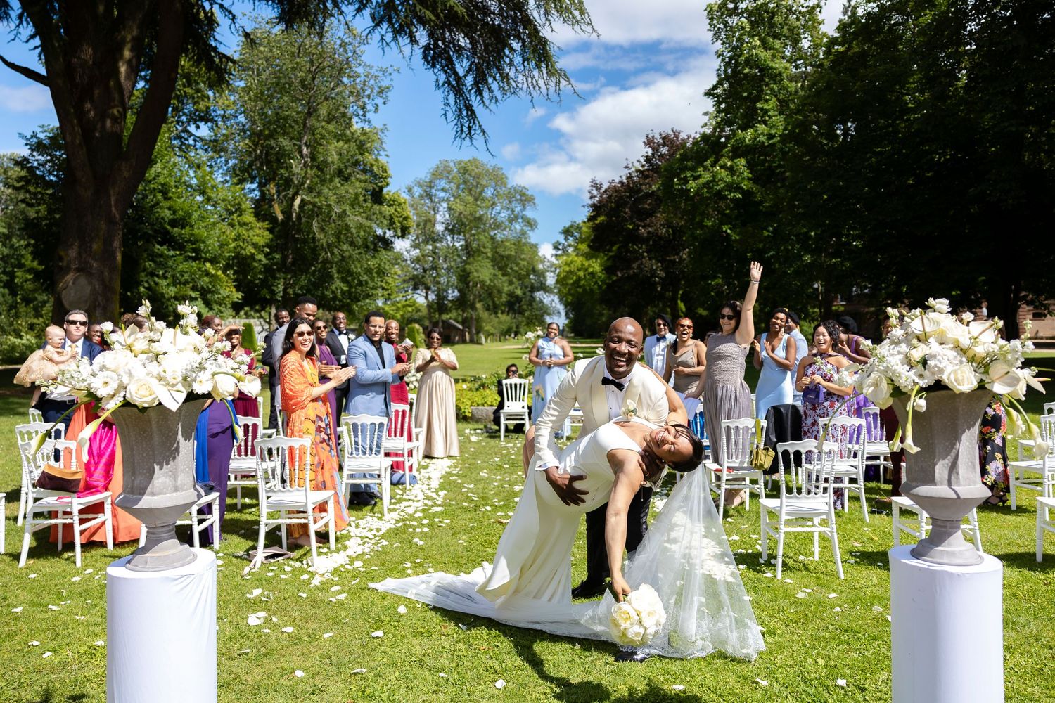 Joyful newlyweds celebrate at outdoor wedding ceremony, groom lifting bride on flower-lined aisle with cheering guests.