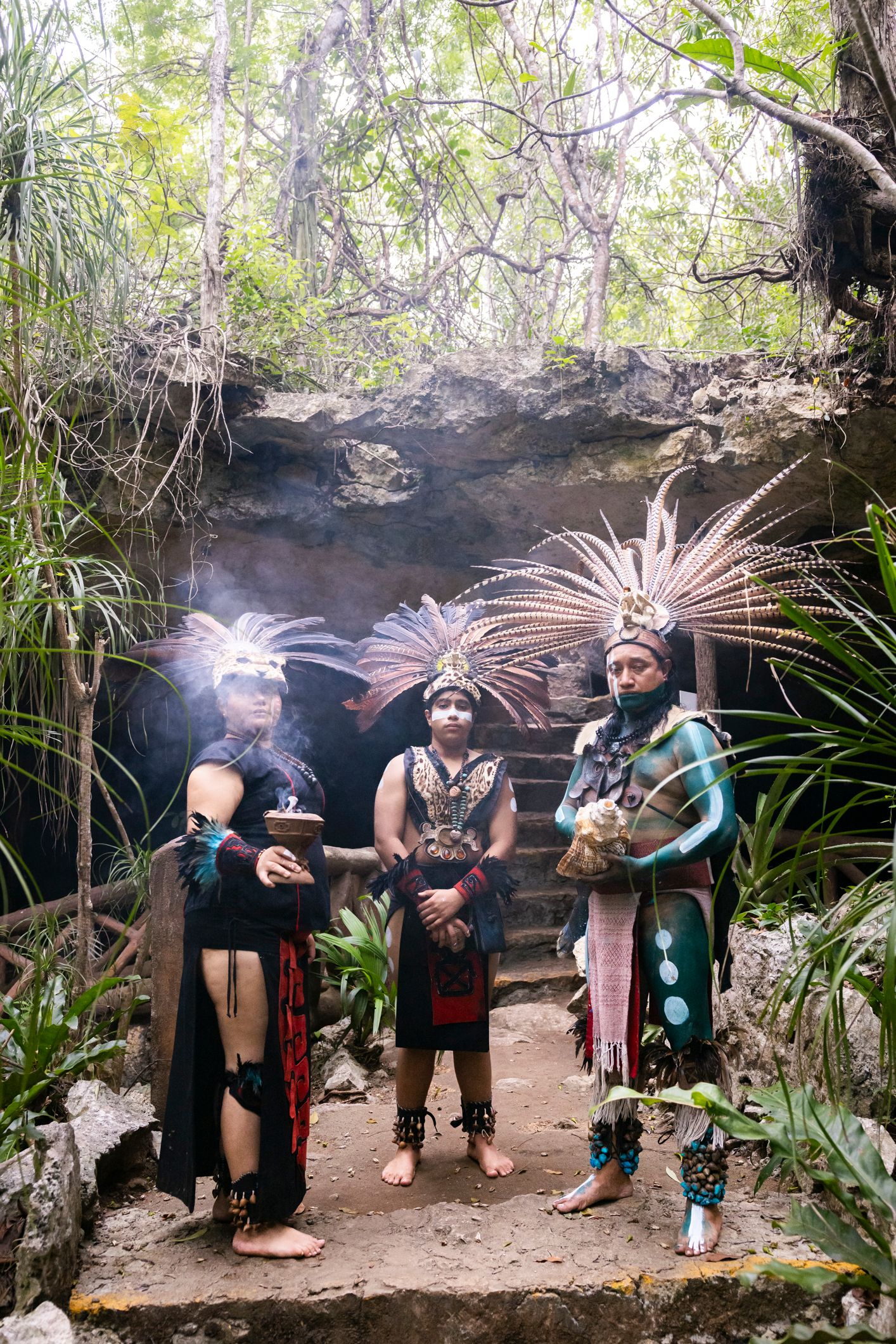 Three performers in traditional Mayan ceremonial costumes with feathered headdresses stand near a jungle cave entrance.