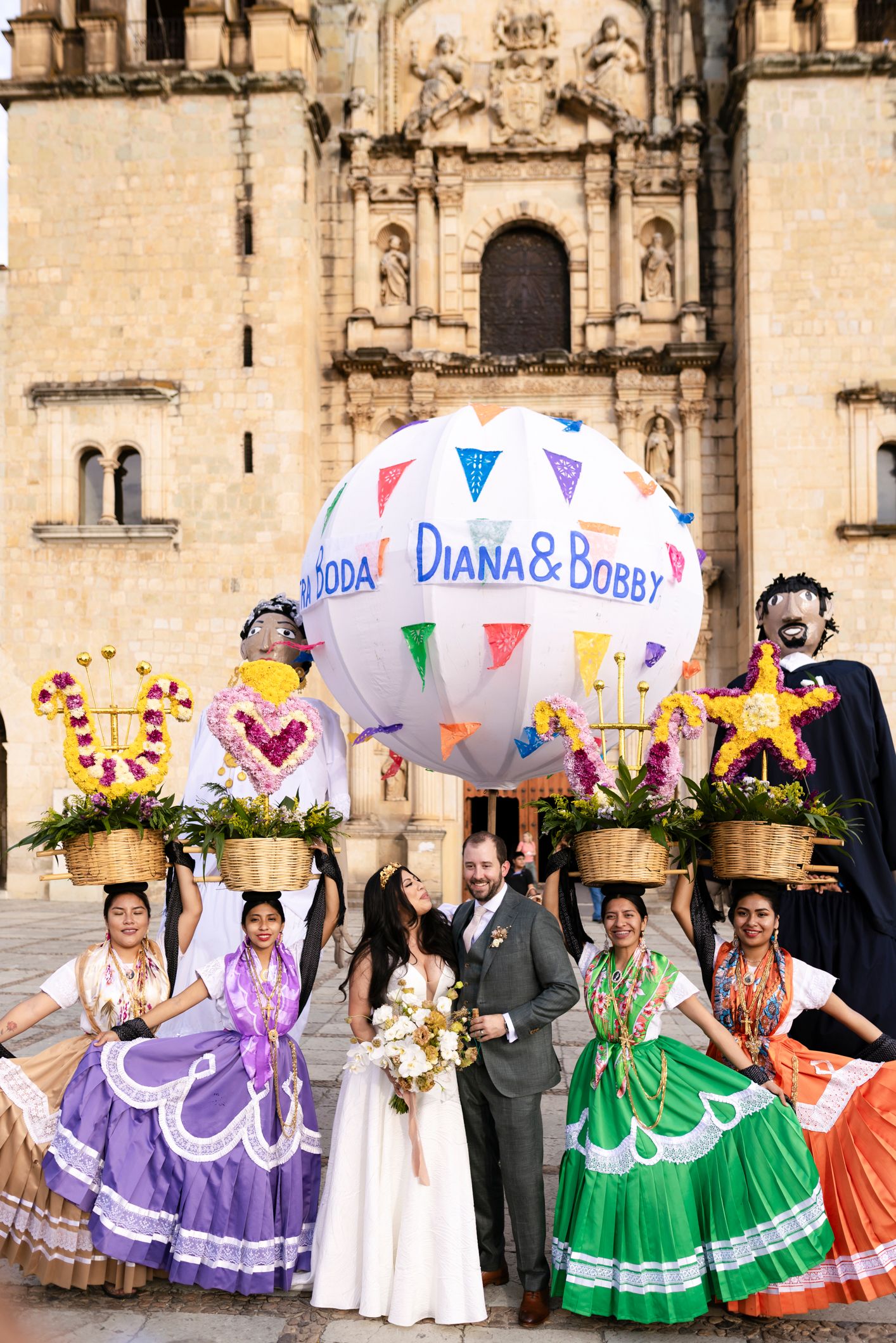 Bride and groom pose with Oaxacan dancers in colorful dresses holding a festive wedding balloon outside a historic church.