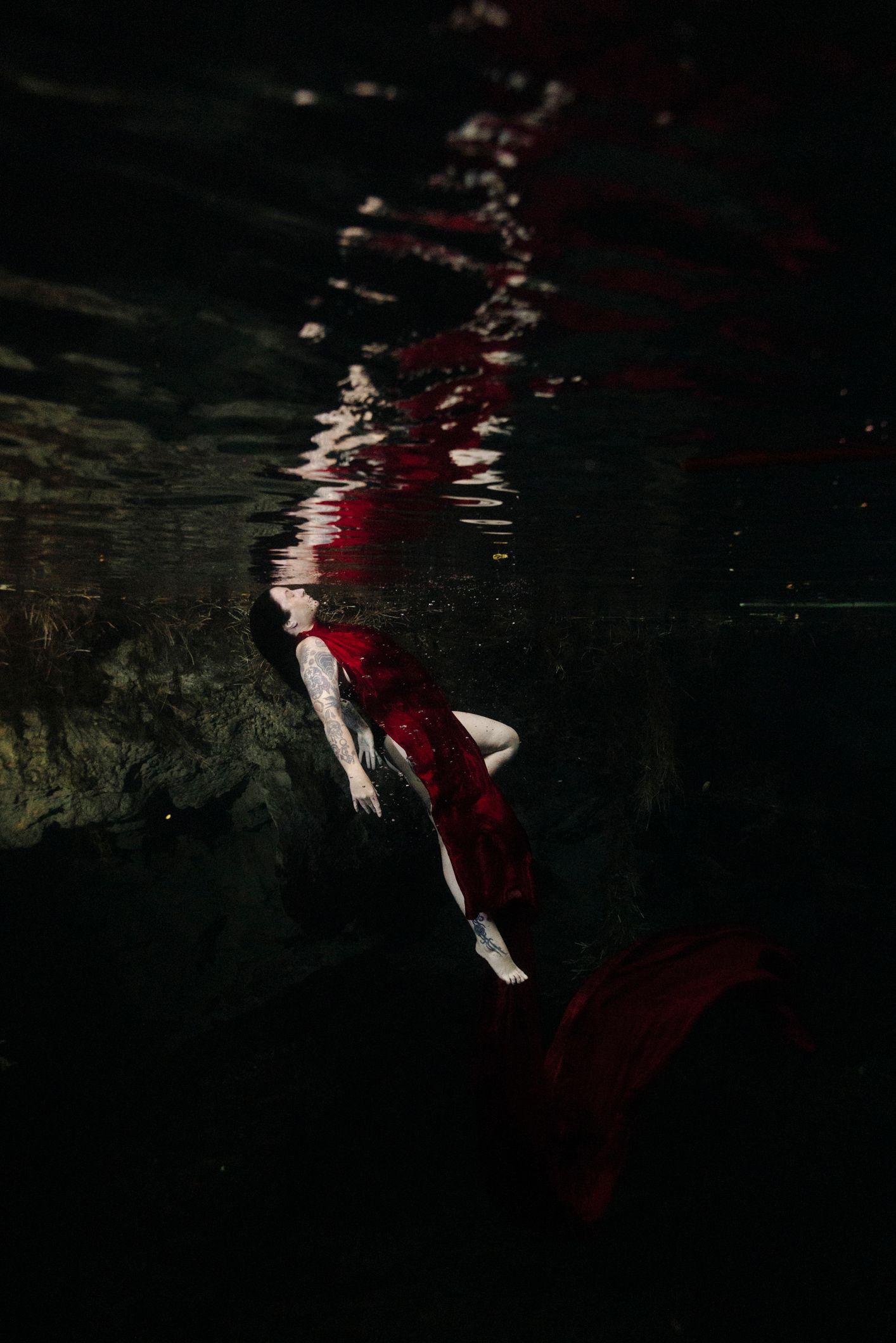 Woman in red dress floating underwater in dark moody pool, artistic fine art photography.