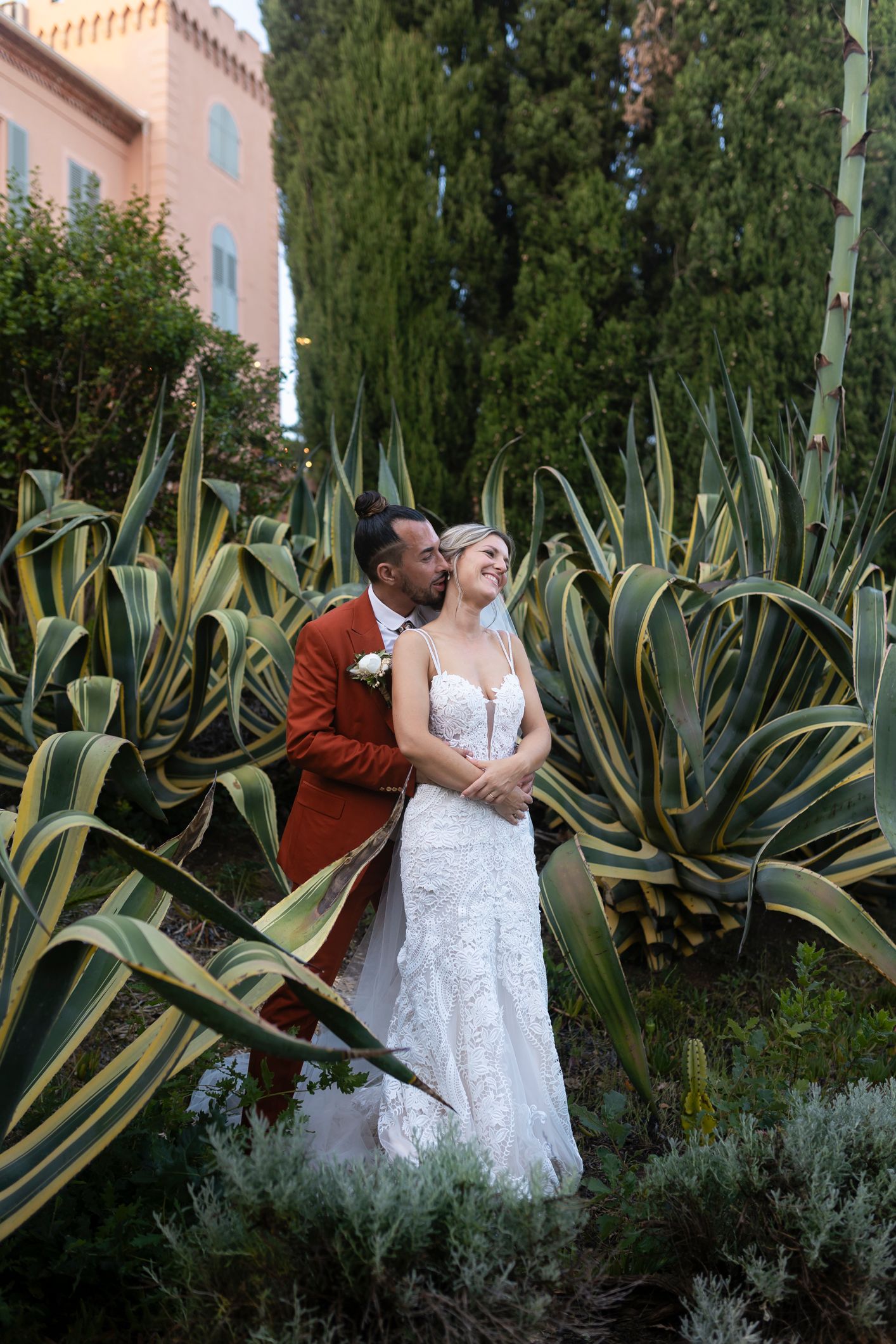 Bride and groom embrace among tall agave plants in a lush garden, groom in rust suit kissing bride in lace wedding gown.
