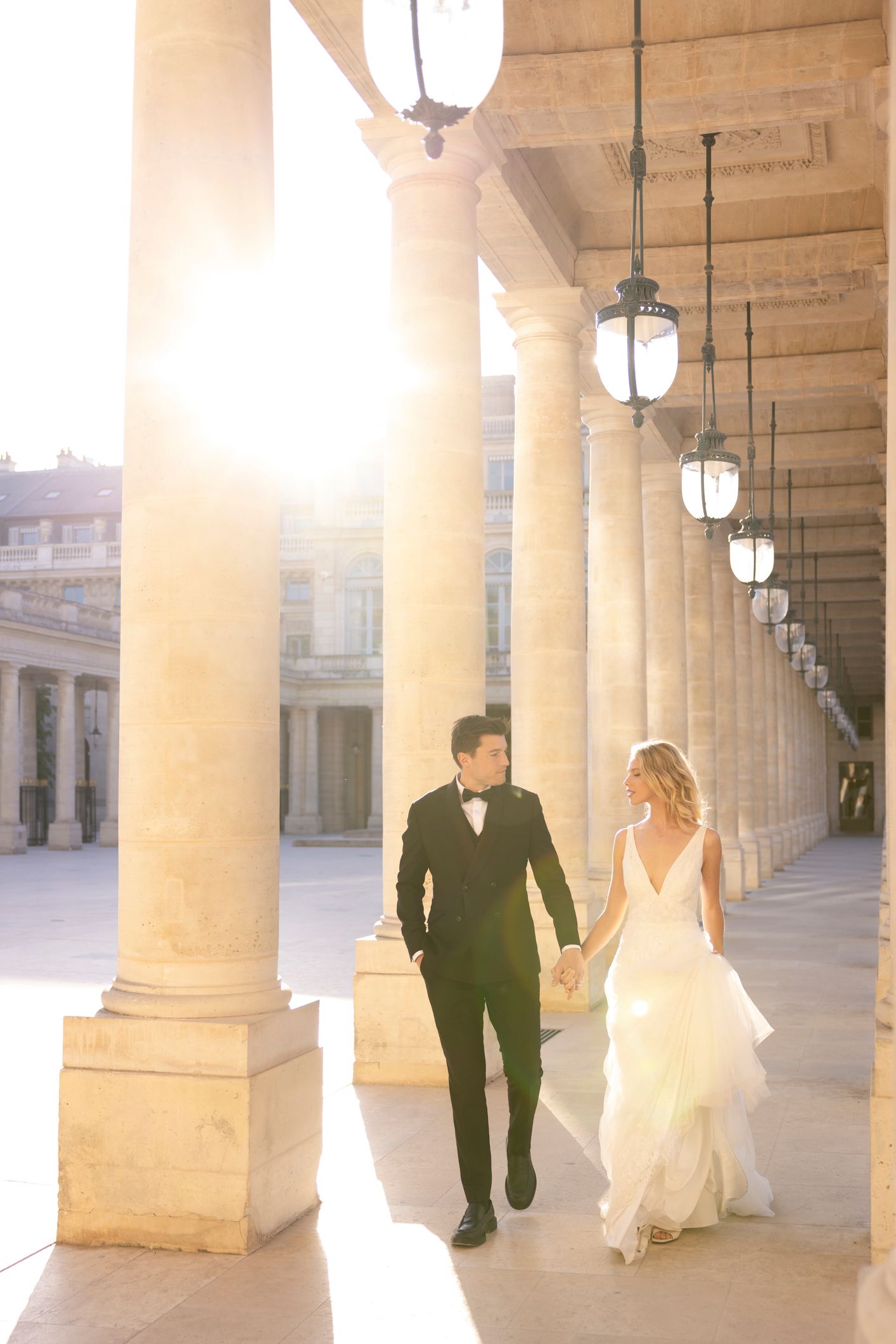 Bride and groom walk hand-in-hand through sunlit Palais Royal colonnade in Paris, wearing elegant wedding attire.