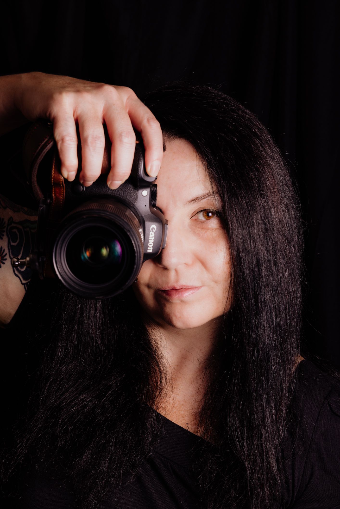 Woman with long dark hair holding a Canon DSLR camera up to her face against a dark background.