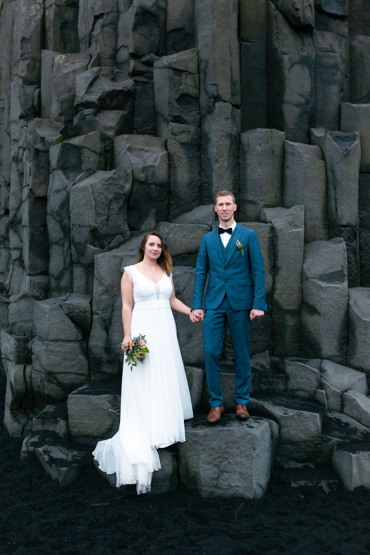 Bride in white gown and groom in blue suit hold hands before dramatic black basalt columns on Iceland's volcanic beach.