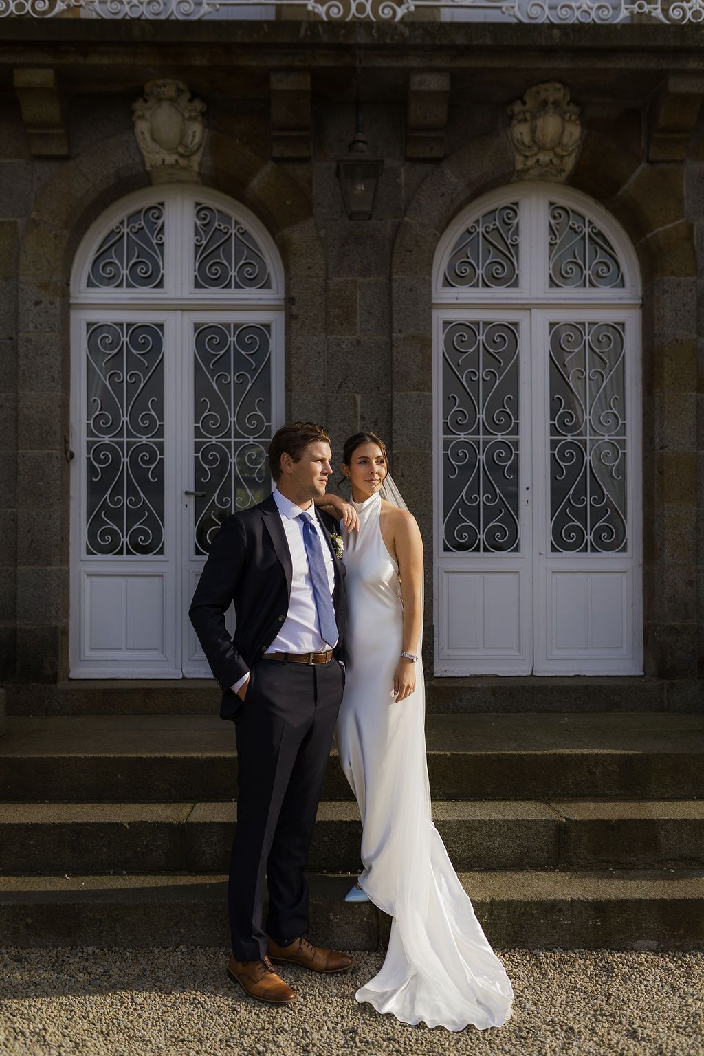 Bride and groom pose elegantly before ornate white doors of a stone building on their wedding day.
