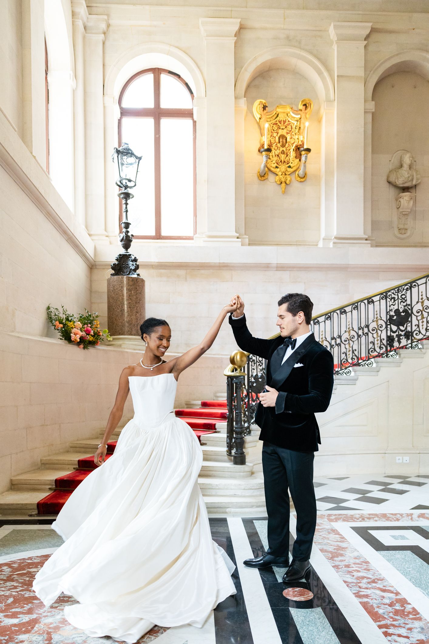 Bride and groom dancing elegantly in a grand marble staircase hall, wearing wedding gown and tuxedo.
