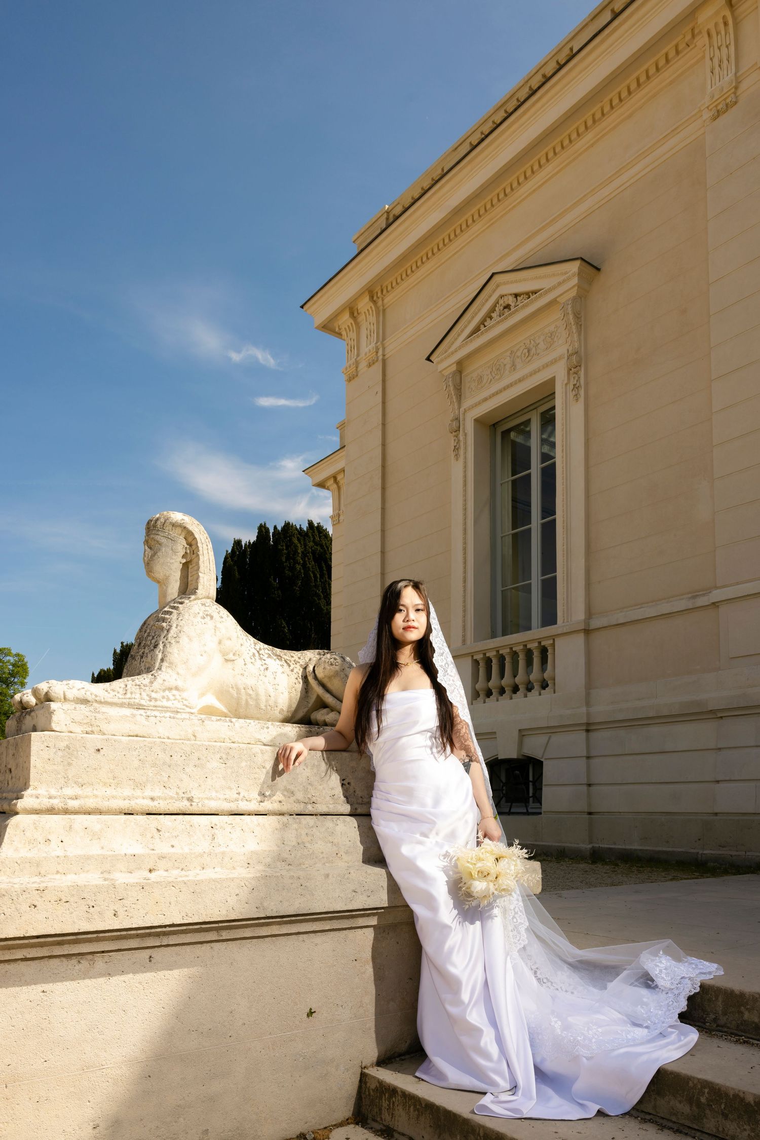 Bride in white gown poses beside stone sphinx sculpture on steps of elegant classical building under blue sky.