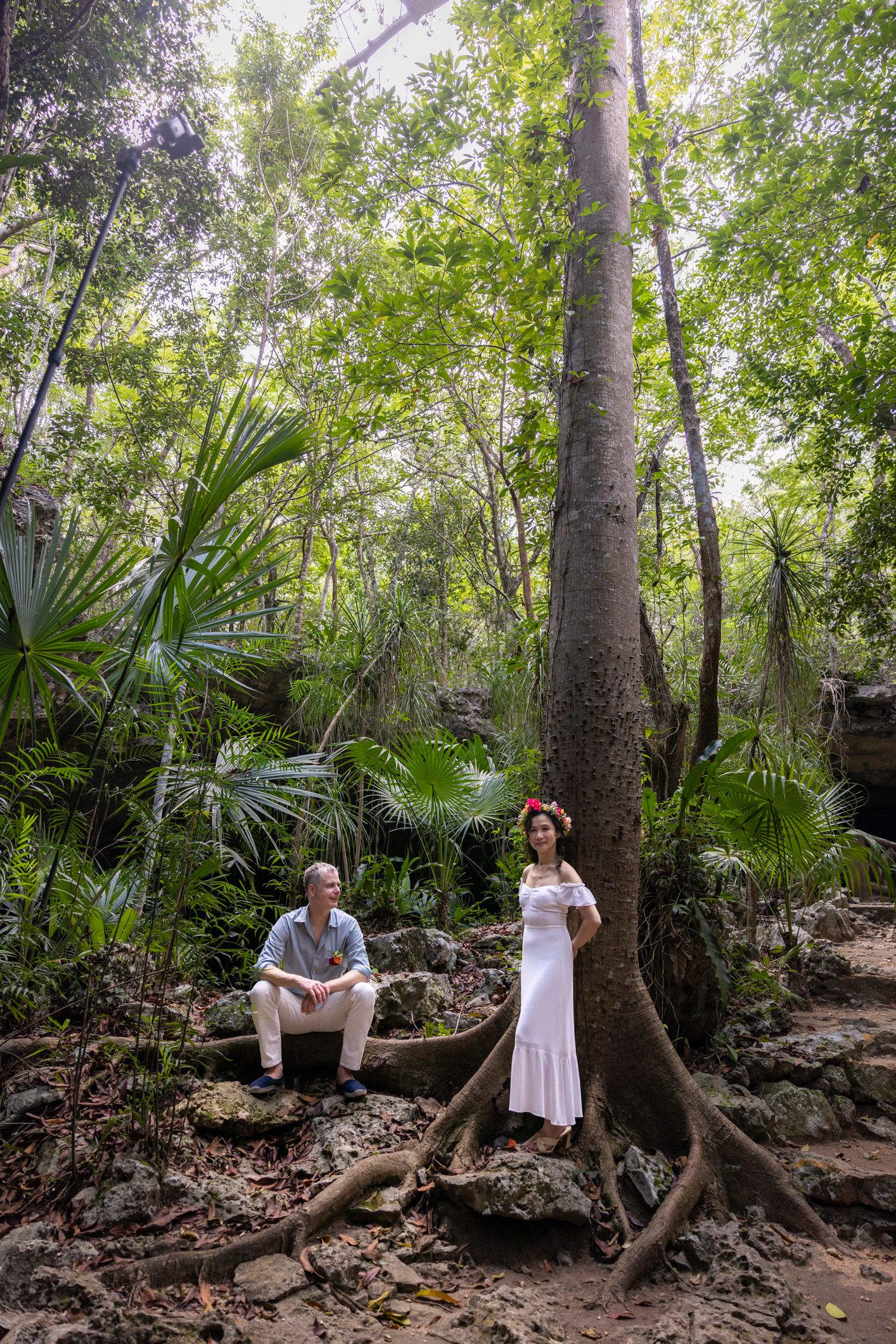 Couple in lush jungle forest, woman in white dress with floral crown stands by large tree, man sits on rocks nearby.