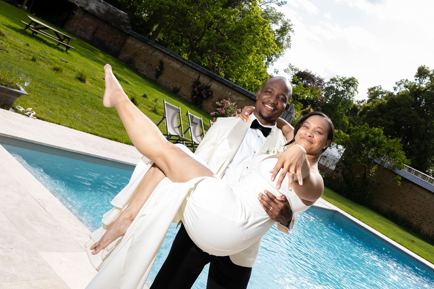 Groom in tuxedo carrying bride in white dress poolside at outdoor wedding venue.