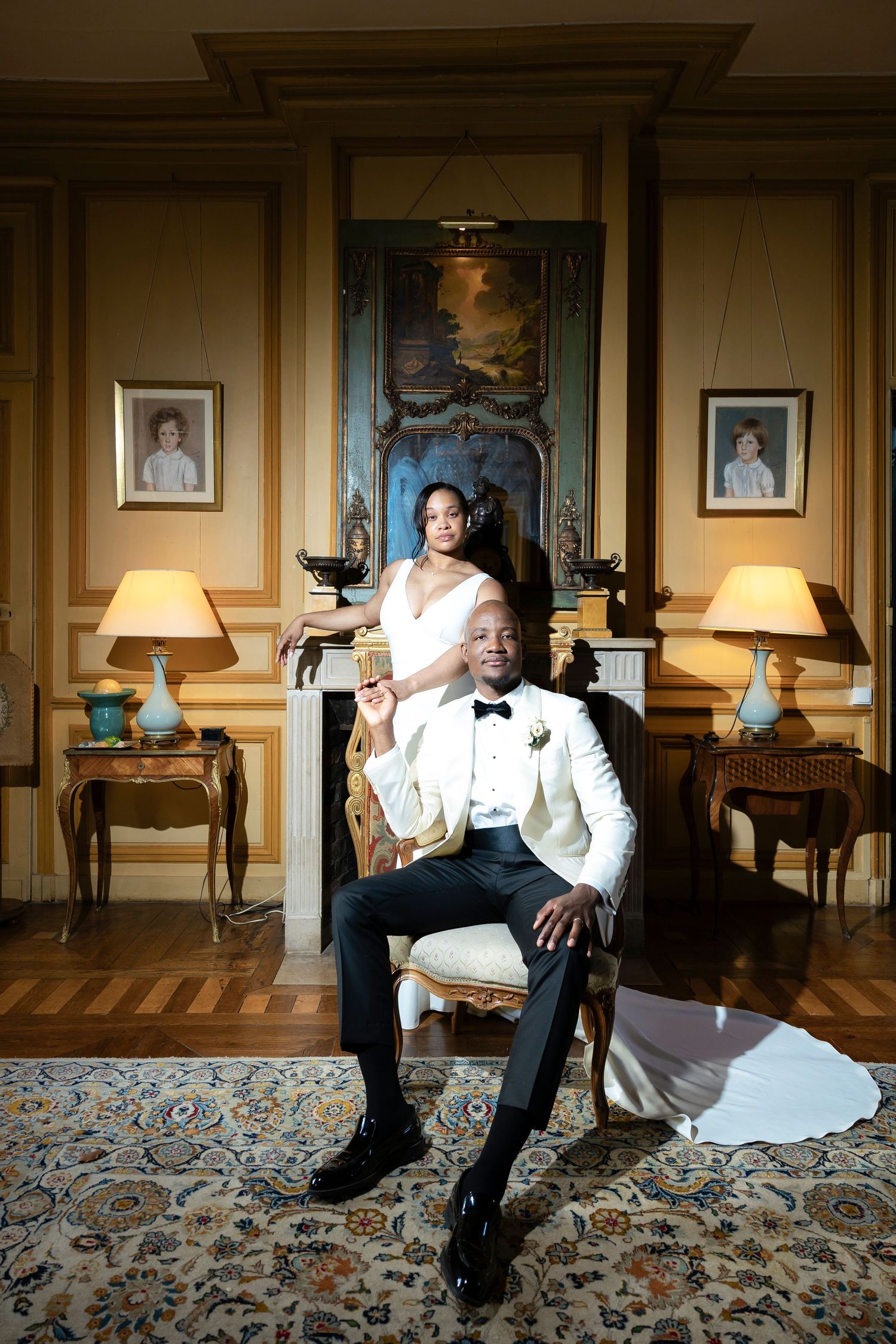 Bride and groom pose in ornate French-style room, groom seated in gilded chair wearing white tuxedo jacket.