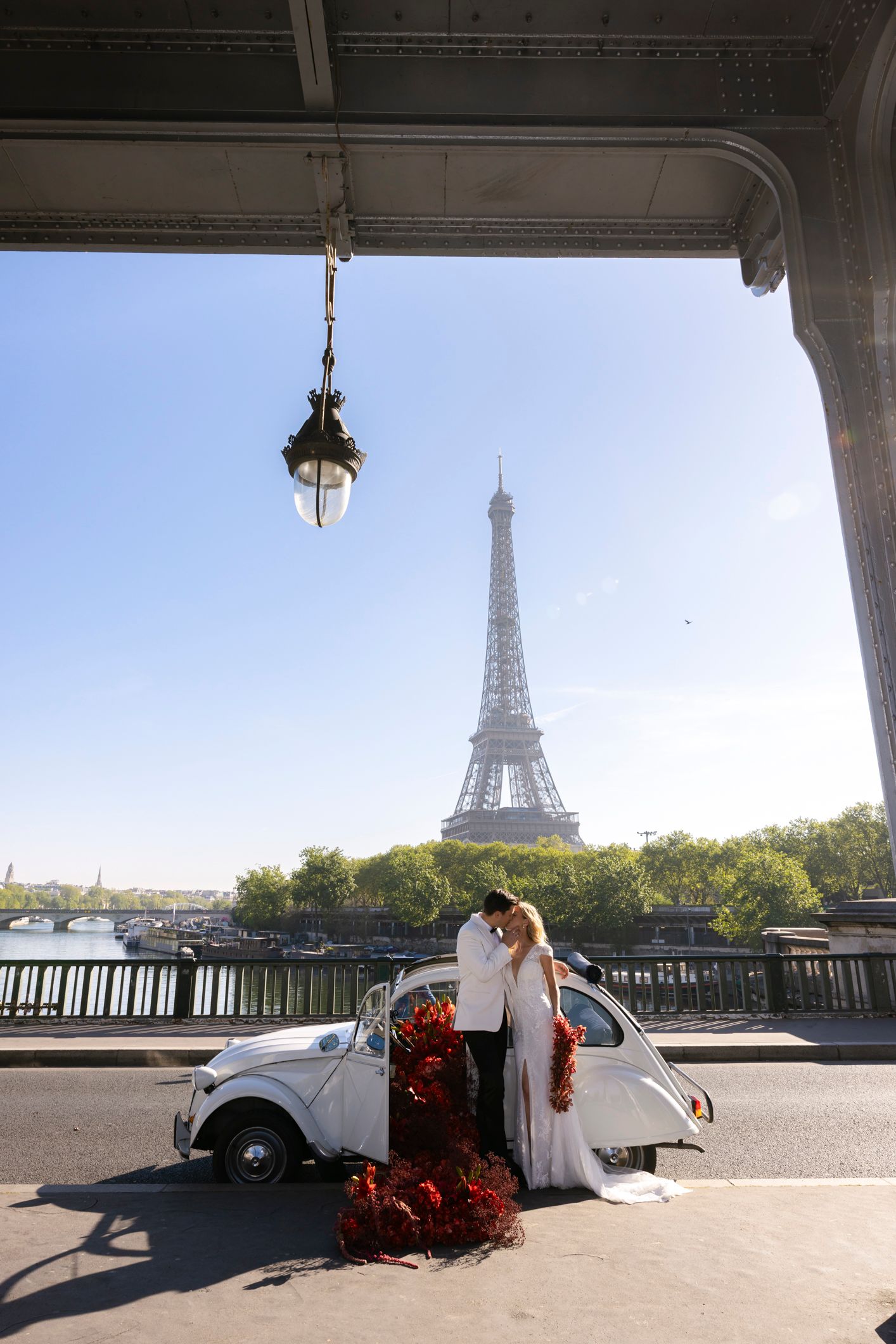 Couple poses by vintage white car on Paris bridge with Eiffel Tower backdrop on sunny day.