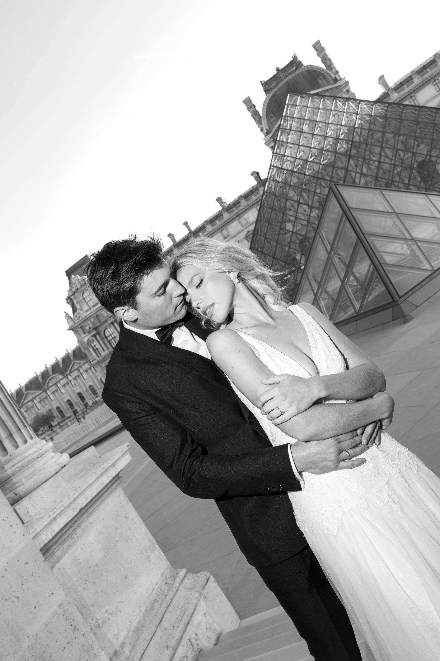 Black and white wedding photo of a couple embracing romantically in front of the Louvre pyramid in Paris.
