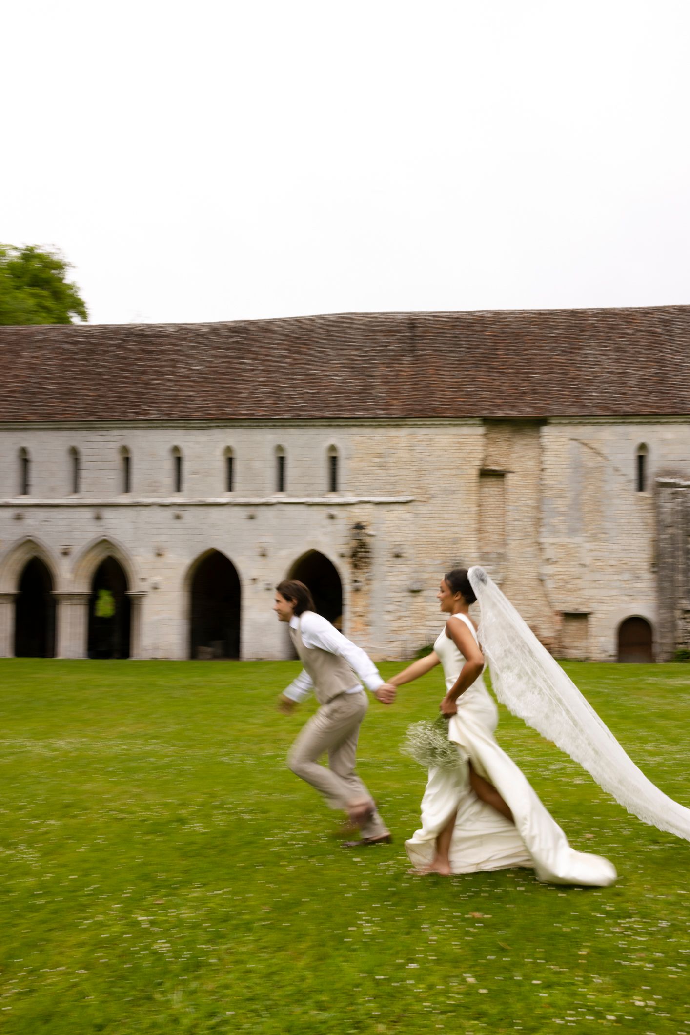 Bride and groom running hand-in-hand across green lawn beside historic stone church on their wedding day.