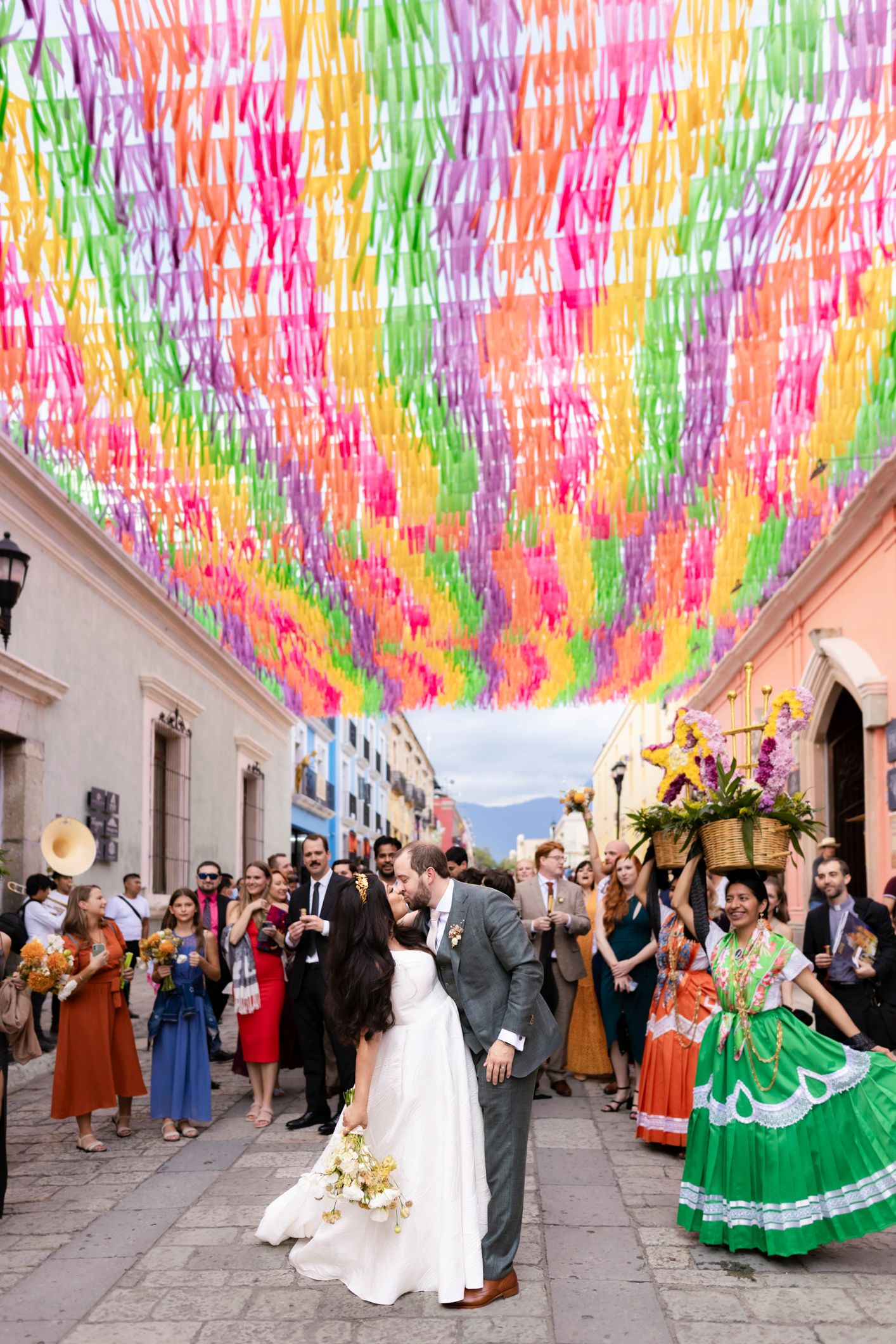 Bride and groom kiss under colorful streamers on a festive Mexican street, surrounded by guests and traditional dancers.