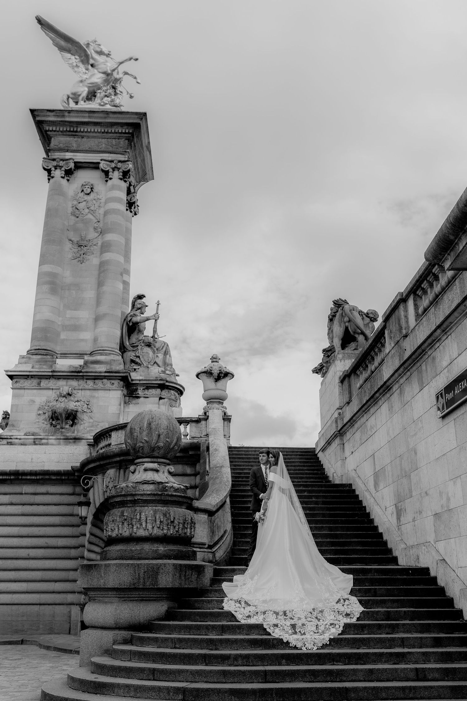 Black and white photo of a bride in a flowing gown and veil on grand stone steps near ornate sculptures.