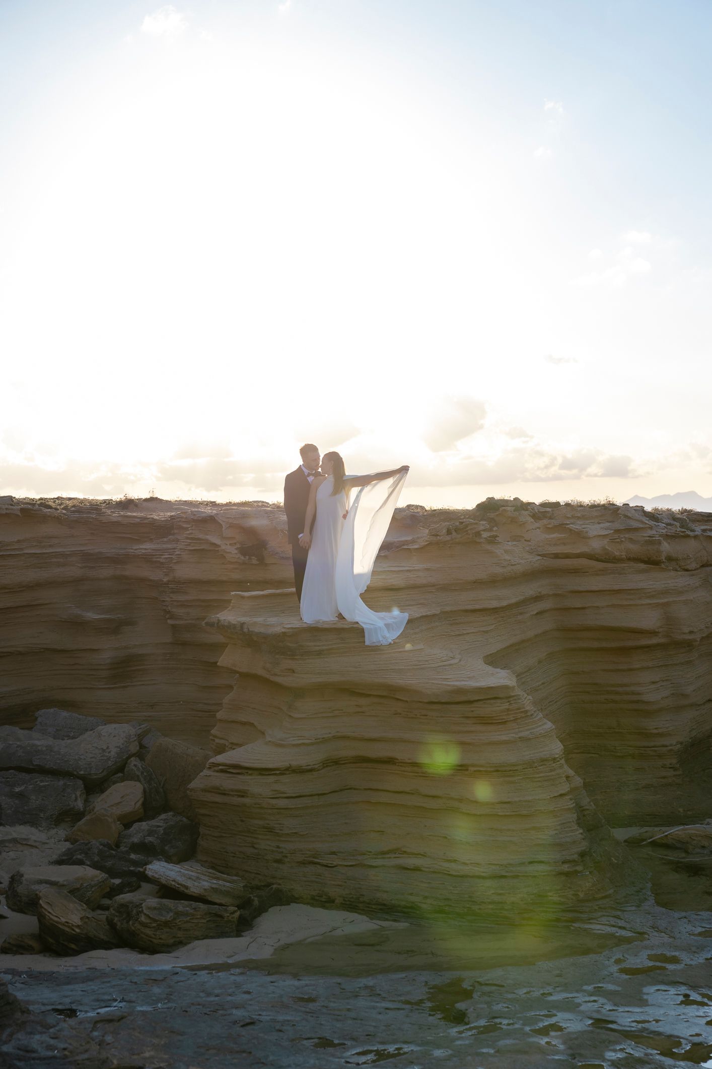 Bride and groom embrace on dramatic rocky cliffs at sunset, wedding veil flowing in the breeze mallorca