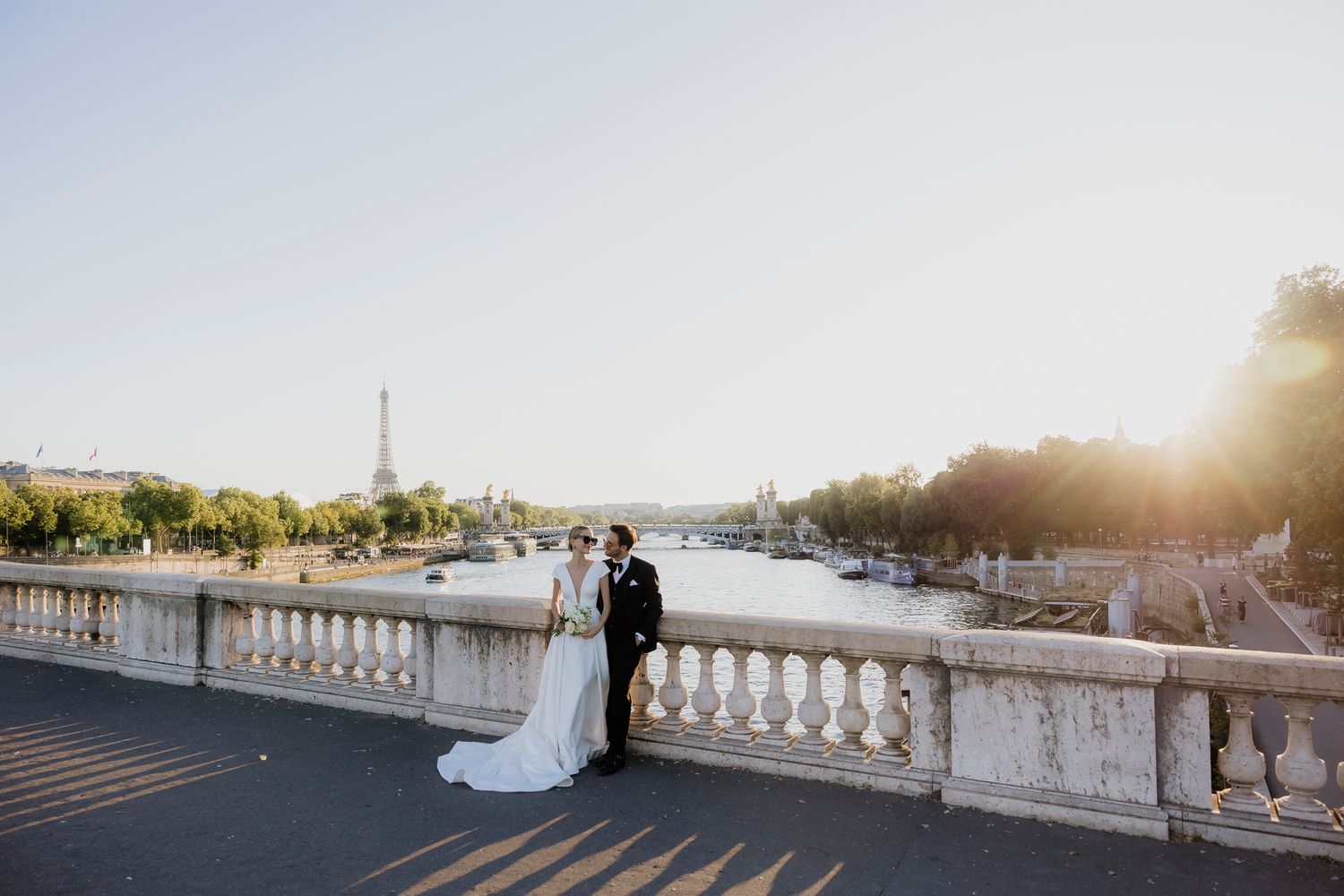 Bride and groom posing on a Parisian bridge over the Seine River with the Eiffel Tower at golden hour.
