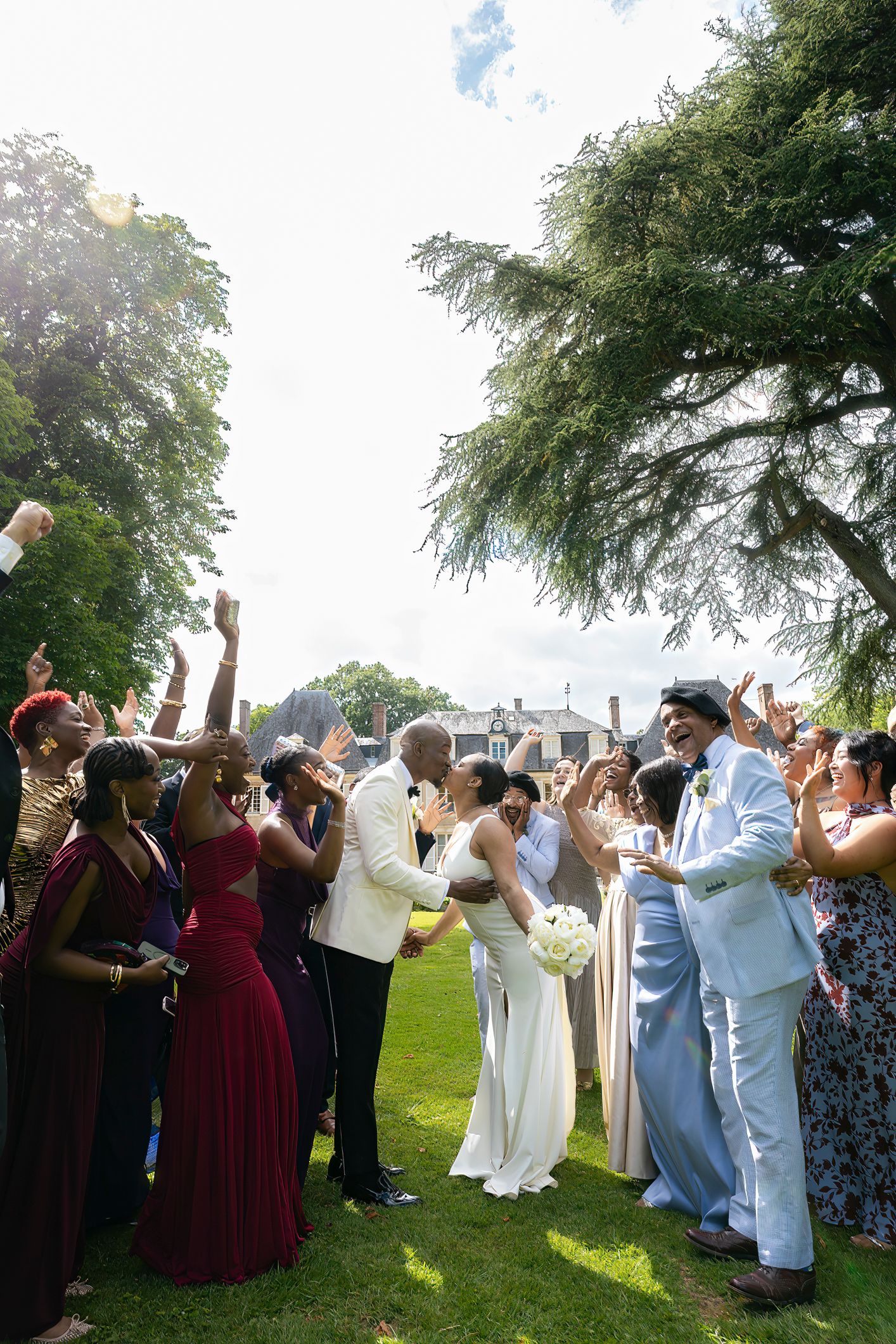 Bride and groom share a kiss surrounded by cheering wedding guests on a sunny outdoor lawn.