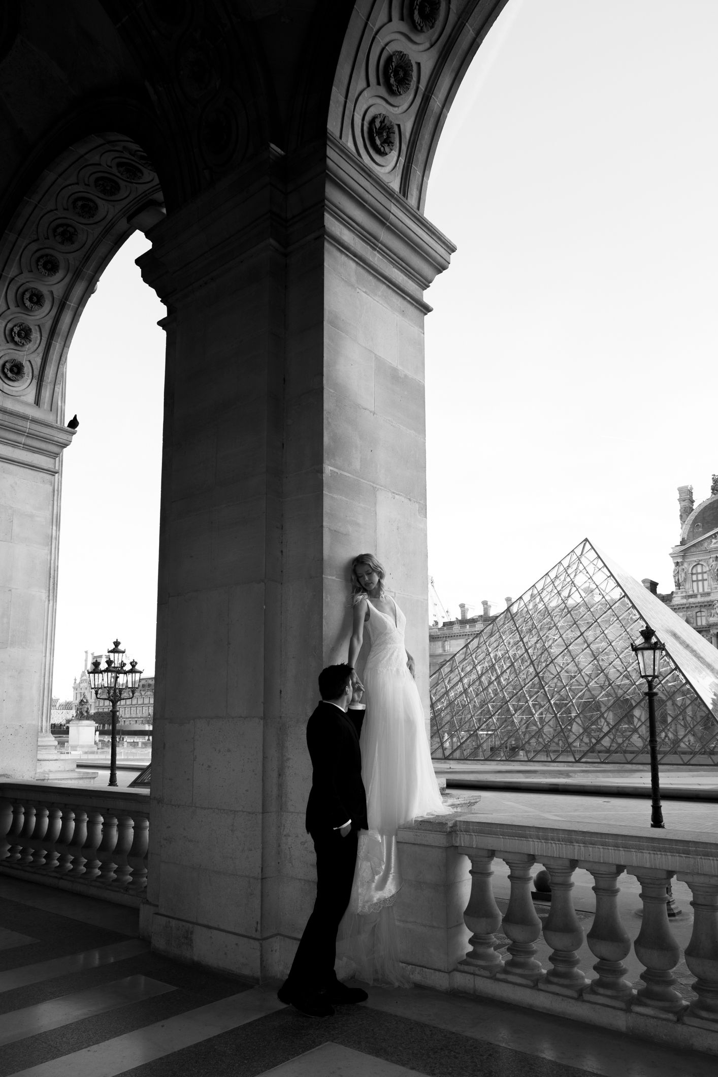 Black and white wedding photo of couple at the Louvre pyramid in Paris, groom lifting bride in elegant gown.