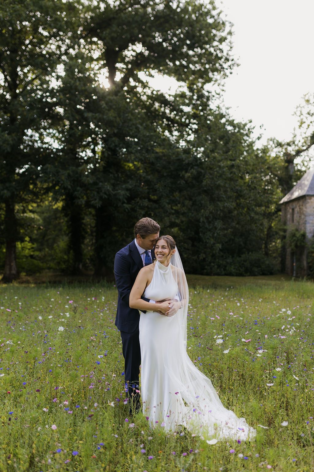 Bride and groom embrace in a wildflower meadow, with lush green trees and golden sunlight in the background.