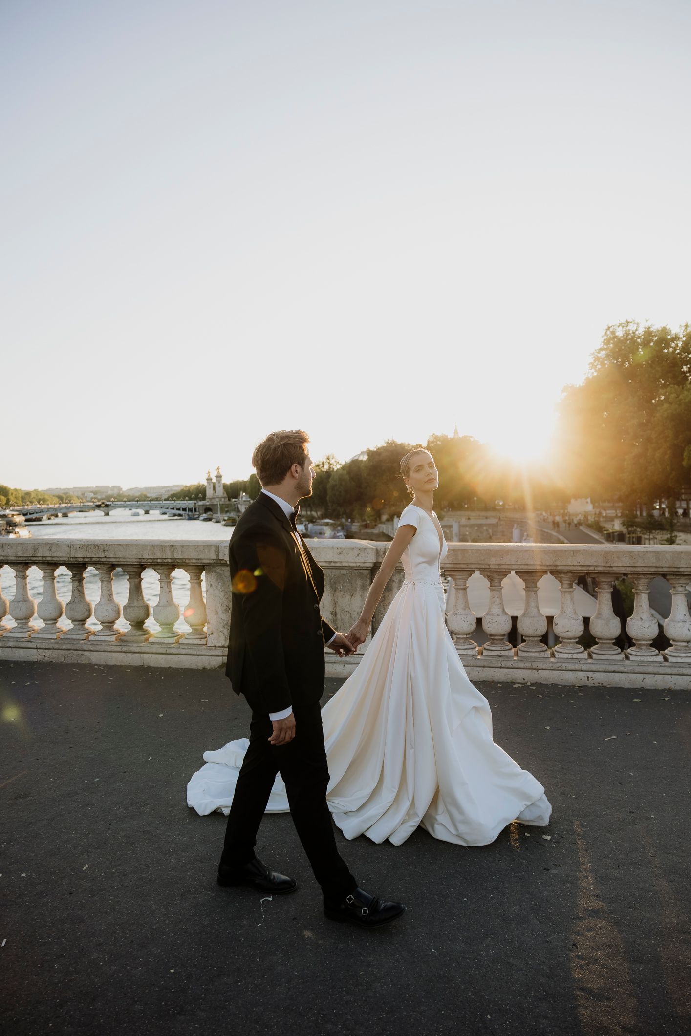 Bride and groom walking hand in hand on a Parisian bridge at golden hour sunset in elegant wedding attire.